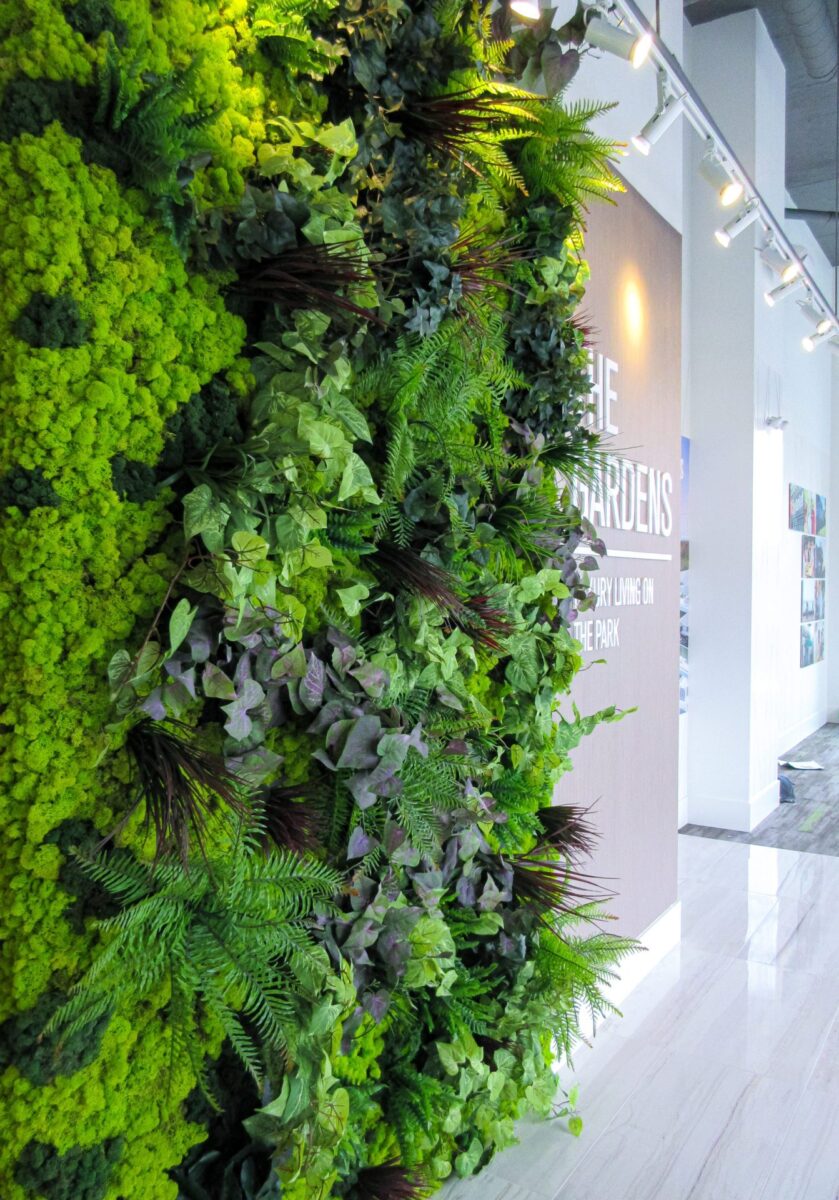 Close-up of an artificial green wall with layered moss, ferns, and mixed foliage, installed beside a building lobby sign.