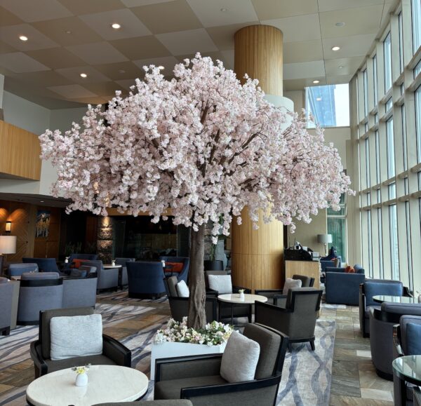 Artificial cherry blossom canopy tree with cascading pink blooms, styled as a statement centerpiece in a hotel lounge with modern seating and floor-to-ceiling windows.