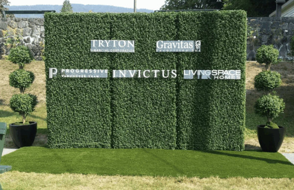 Artificial boxwood hedge backdrop with sponsor logos, flanked by potted topiary trees on grass.