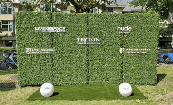 Artificial boxwood hedge backdrop with corporate logos and branded balls on grass for an outdoor event.