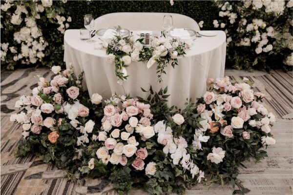 Blush pink and white floral head table arrangement with roses, orchids, and greenery for a romantic wedding design.