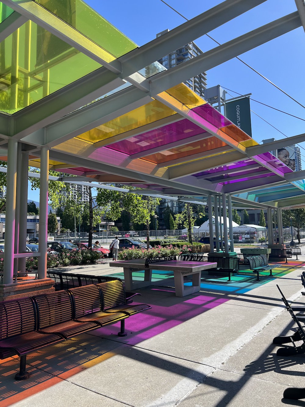 Brightly coloured rainbow decals on glass awnings cast rainbow shadows on summer patio furniture at Coquitlam Centre