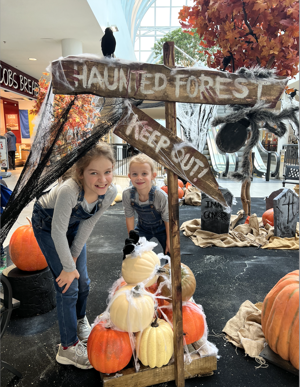 Two kids exploring Greenscape Design's Halloween decor at Lougheed Town Centre in Vancouver, surrounded by artificial gold, white, and orange pumpkins with tall red maple tree, creating a haunted forest ambiance.