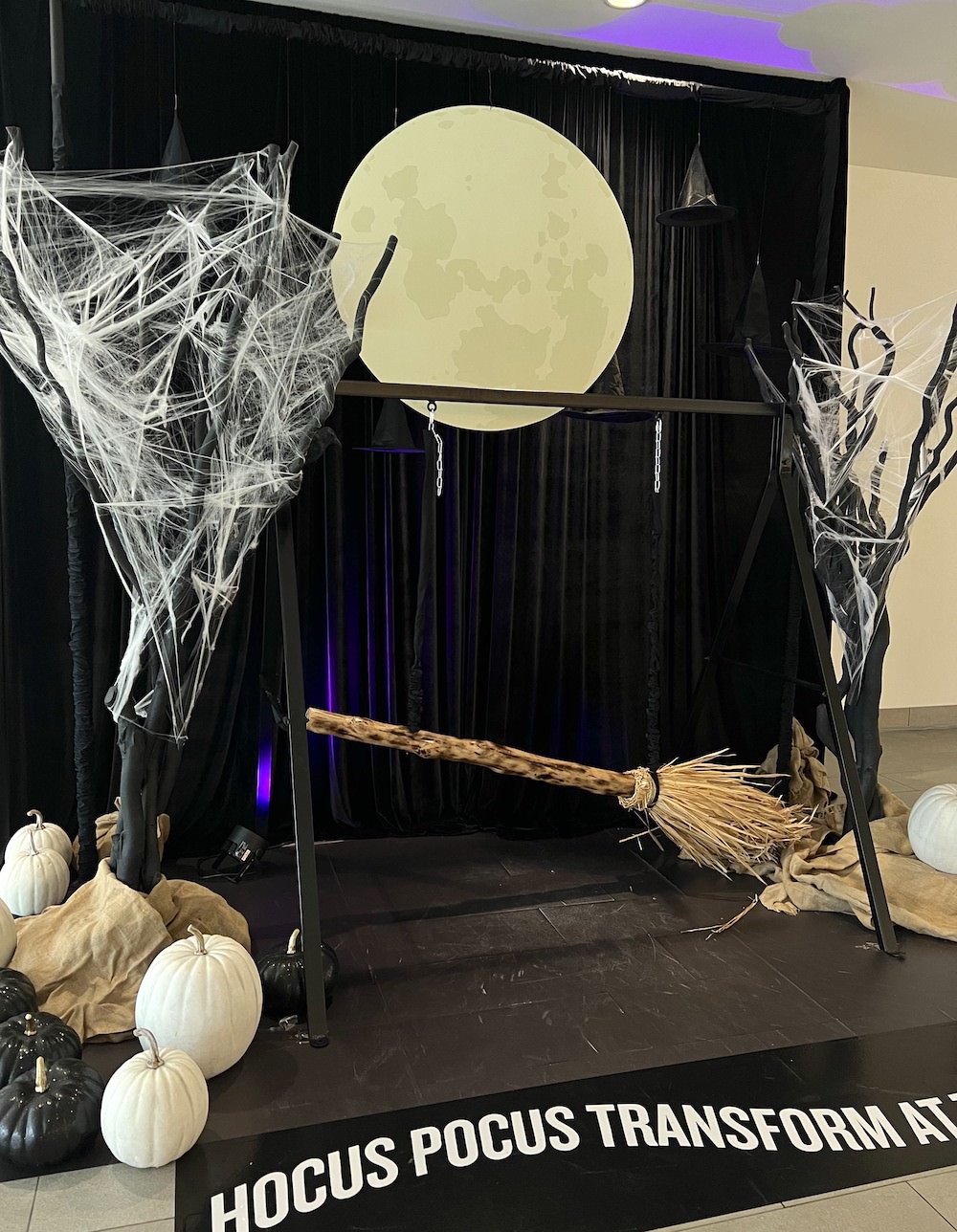 Prop witches' brooms for guests to sit on frame with spooky black trees, cobwebs, black and white pumpkins against a black backdrop, and oversize moon. Greenscape Design's Halloween display at Lougheed Town Centre.