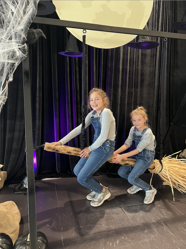 Two kids posing on prop witches' brooms amid spooky black trees, cobwebs, black and white pumpkins against a black backdrop, and an oversize moon. Greenscape Design's Halloween display at Lougheed Town Centre.
