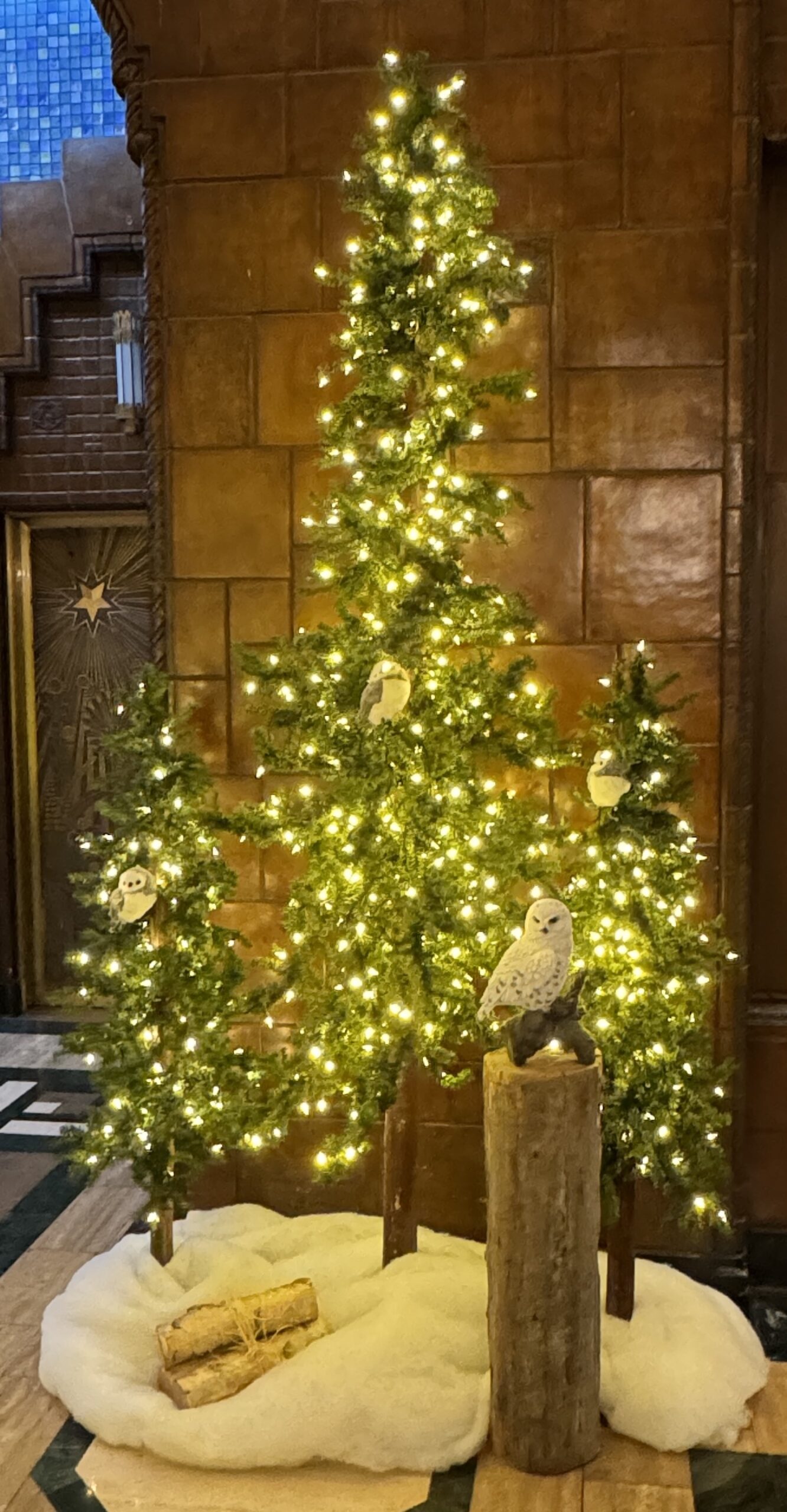 Artificial alpine tree display with warm white lights, snowy base, and owl decorations in an office lobby, creating a winter wonderland ambiance.