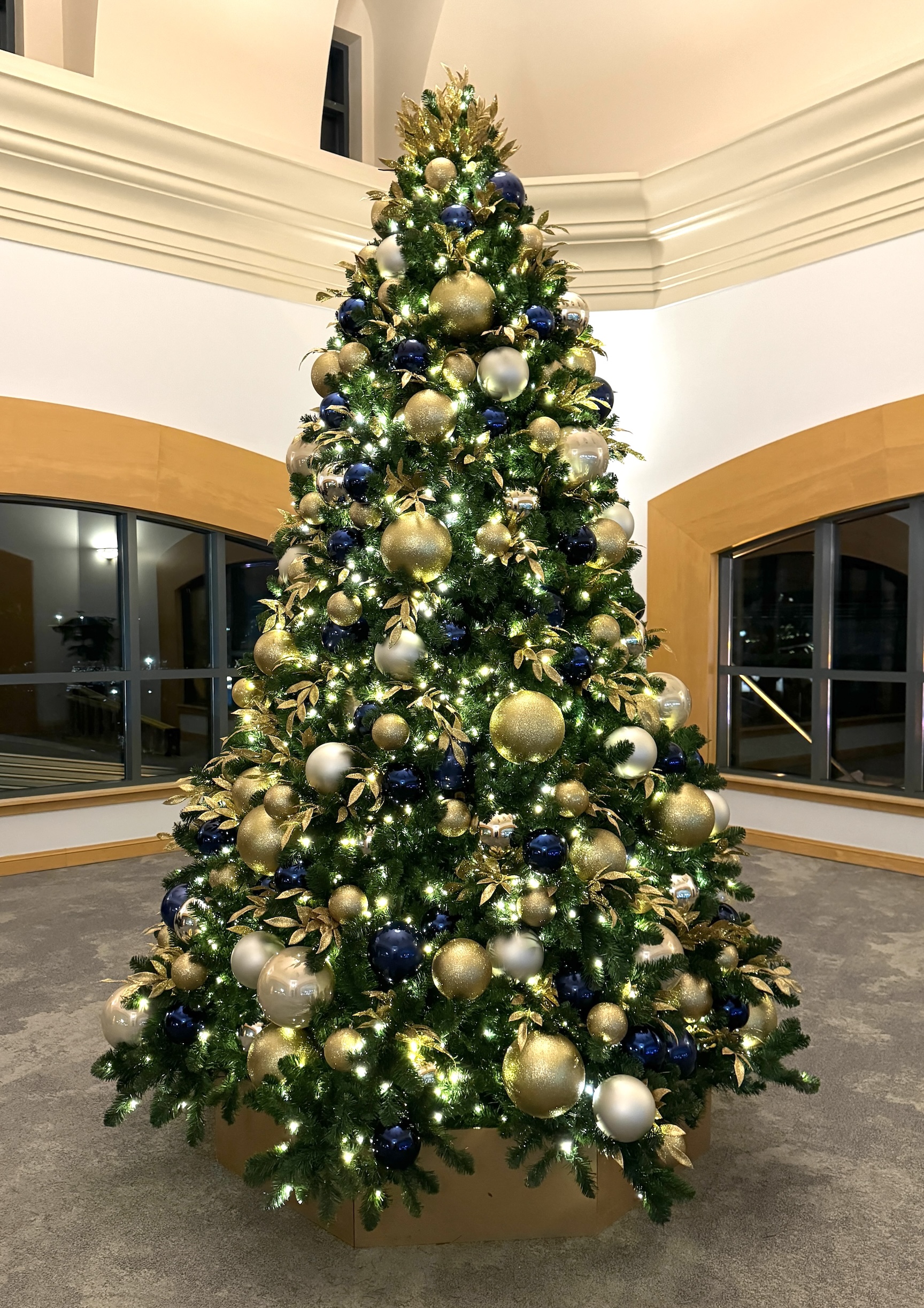 Tall artificial Christmas tree adorned with gold and navy ornaments, warm lights, and lush greenery, displayed in a hotel atrium.