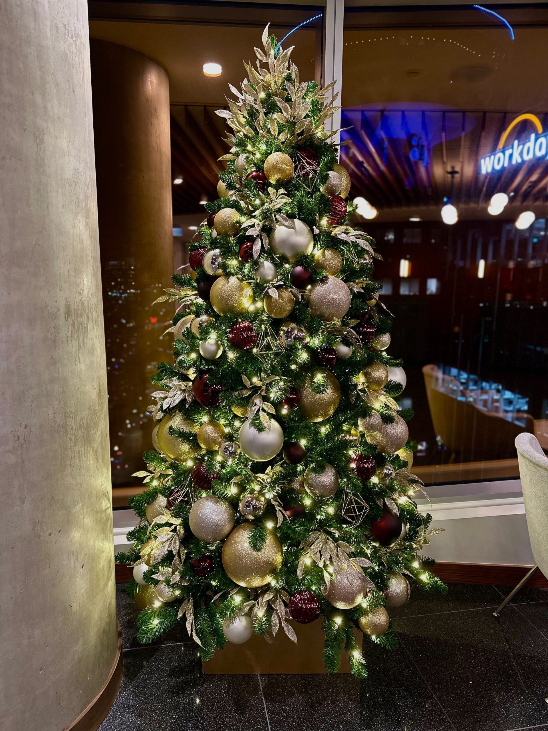 Luxurious Christmas tree adorned with burgundy, gold, and oat decorations, complemented by warm white lights and metallic ornaments, displayed in a modern indoor setting.