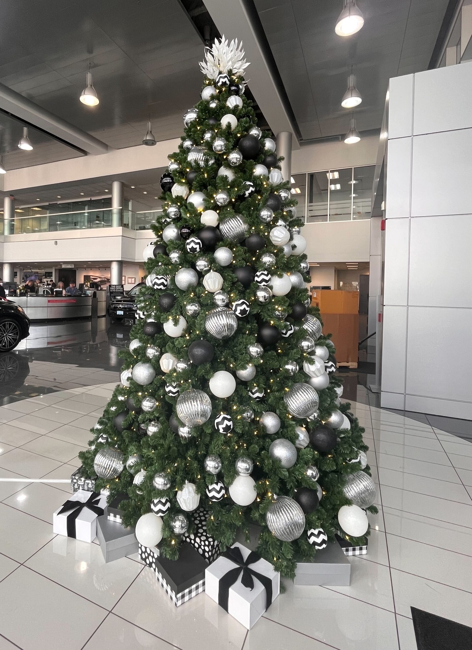 Christmas tree decorated with black, white, and silver ornaments, surrounded by matching gift boxes at a car dealership.