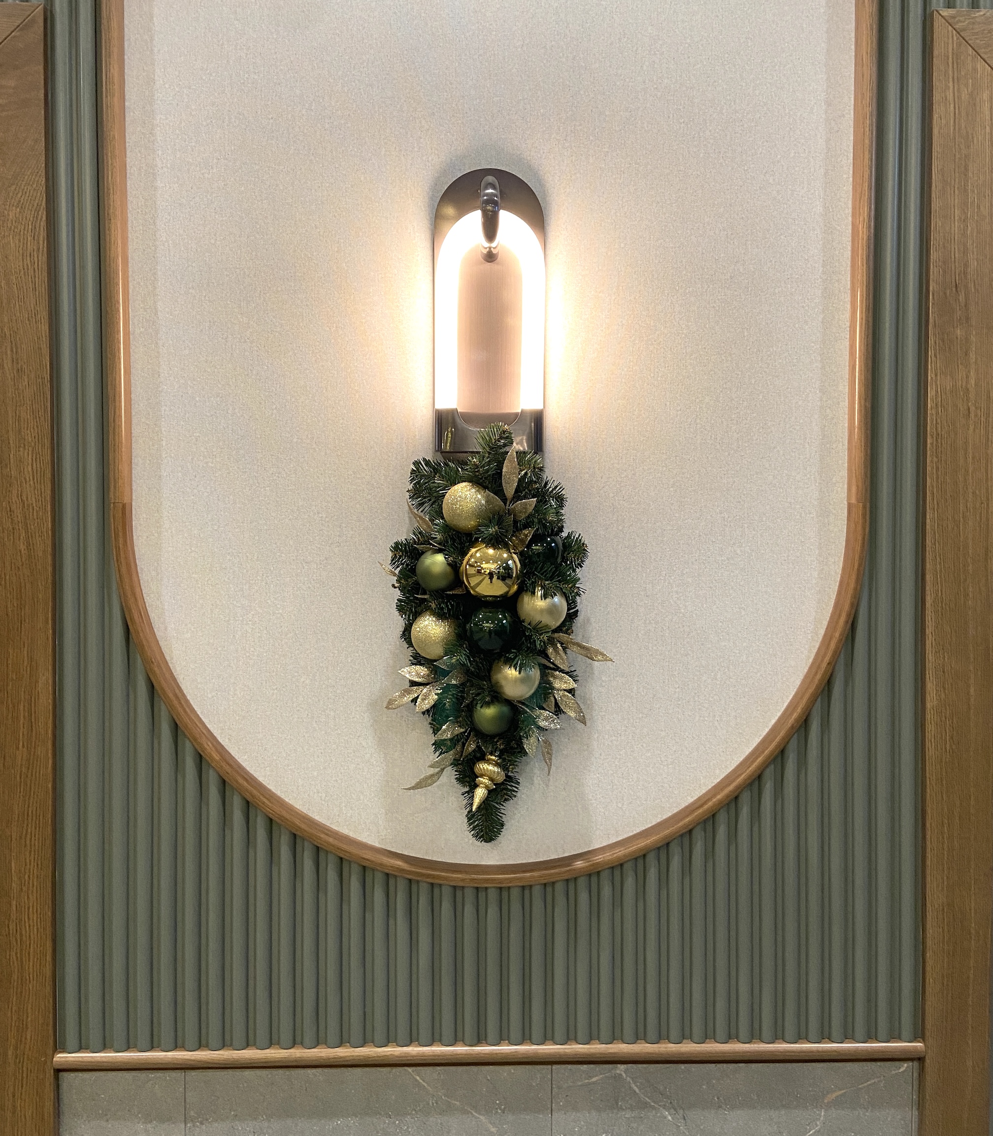 Festive Christmas swag featuring green and gold ornaments with lush greenery and golden accents, beautifully mounted beneath a modern sconce.