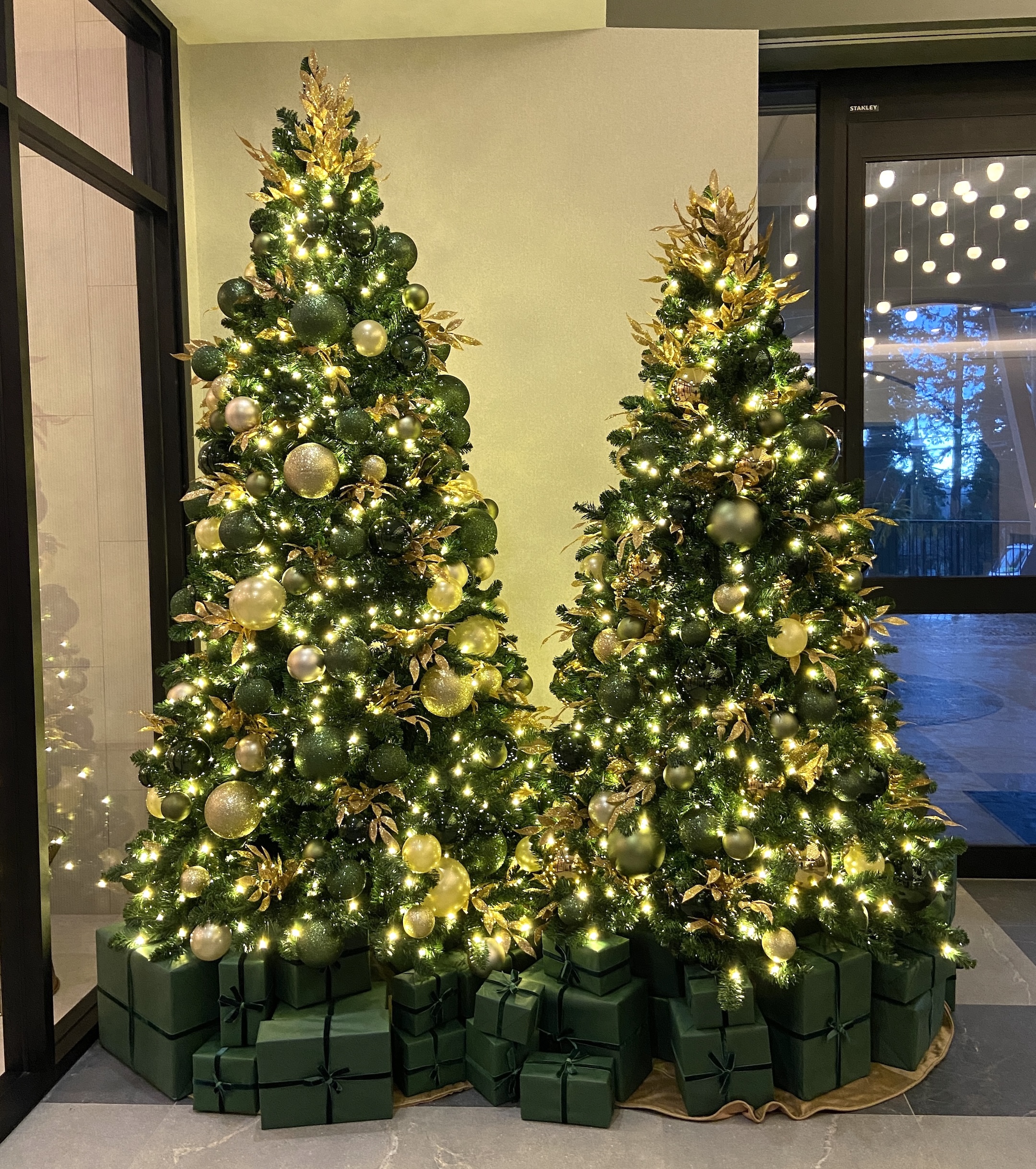 Two elegant Christmas trees decorated with green and gold ornaments, warm lights, and matching gift boxes, displayed in a modern gold resort setting.