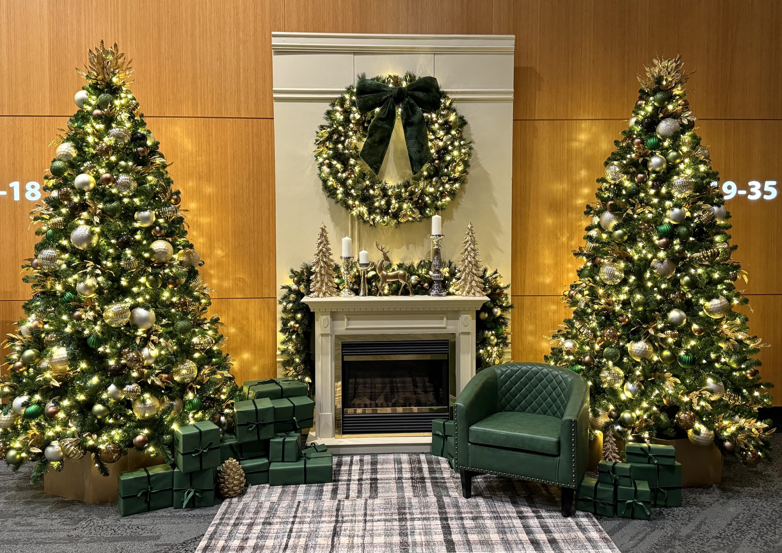 Christmas setup with two decorated green and gold trees, a wreath with a velvet bow, a fireplace mantle adorned with candles, and matching wrapped gift boxes.