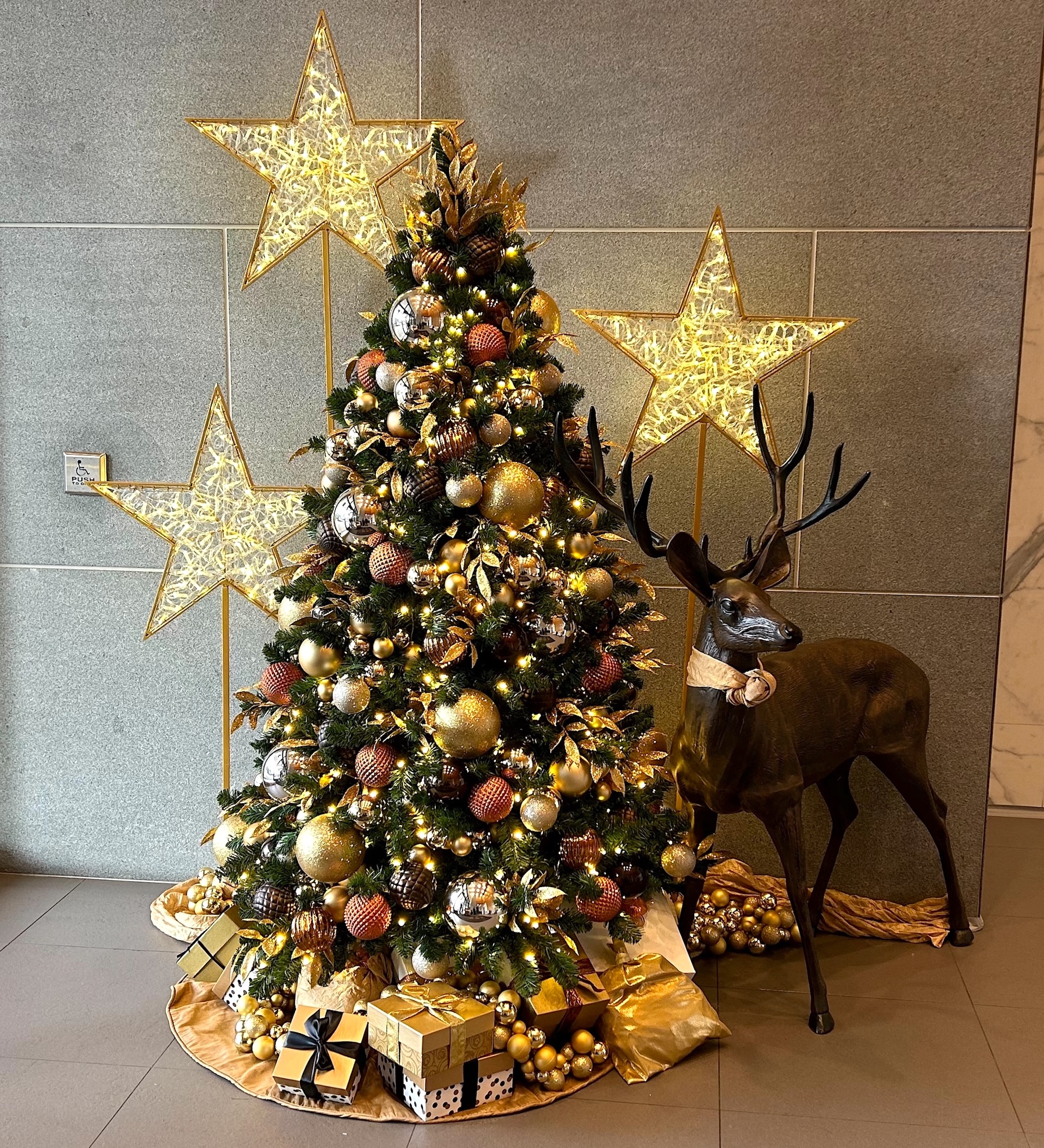 Holiday tree decorated with gold, bronze, and copper ornaments, surrounded by illuminated stars, gift boxes, and a decorative reindeer in a modern setting.