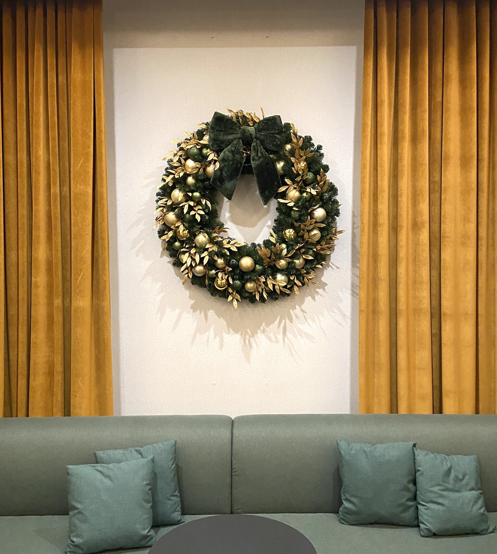 Luxurious Christmas wreath adorned with gold ornaments, greenery, and a green velvet bow, displayed against a modern backdrop with golden curtains in a hotel lobby.