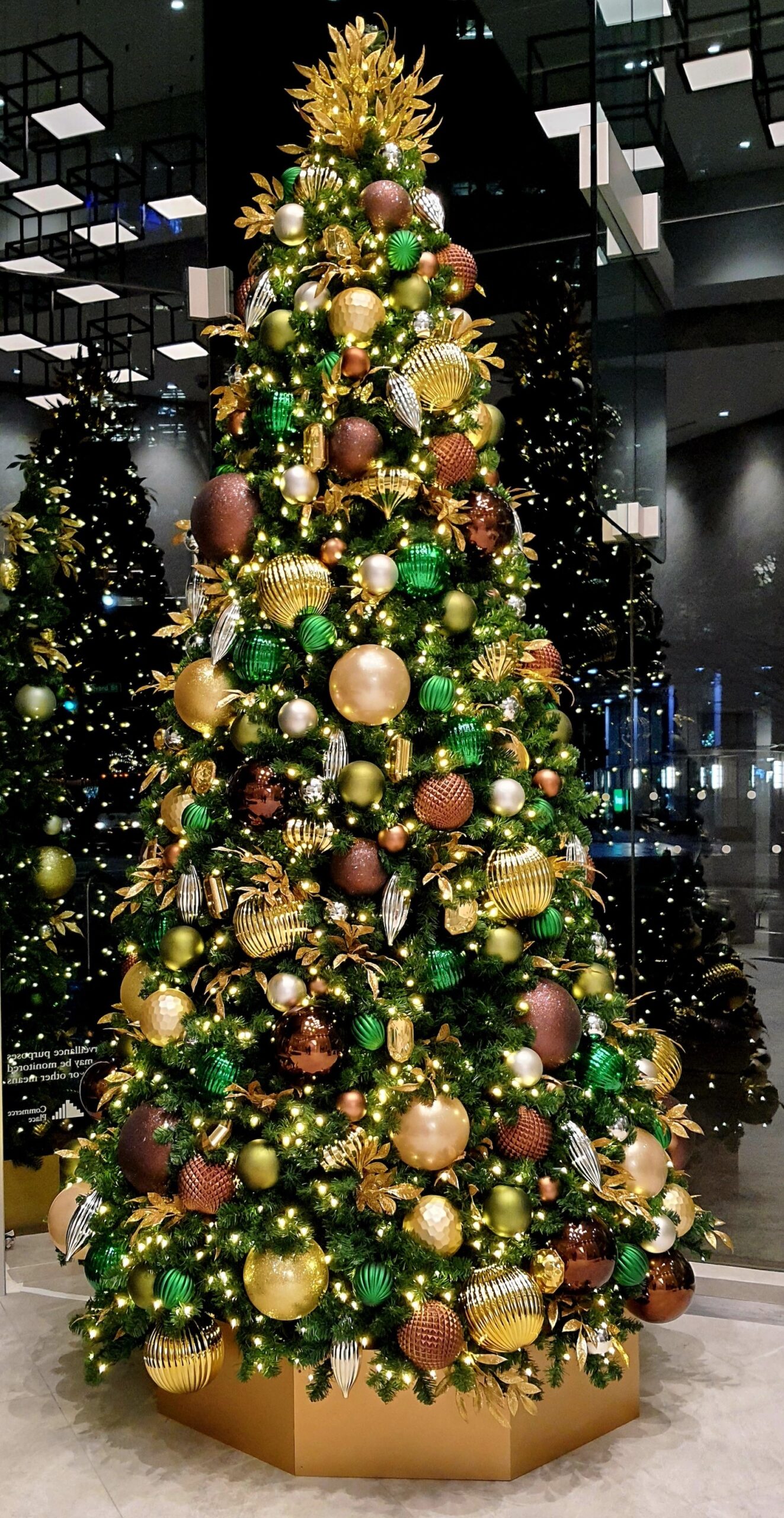Christmas tree adorned with green, gold, and bronze ornaments, featuring intricate textures and warm lights, displayed in a Vancouver office lobby.