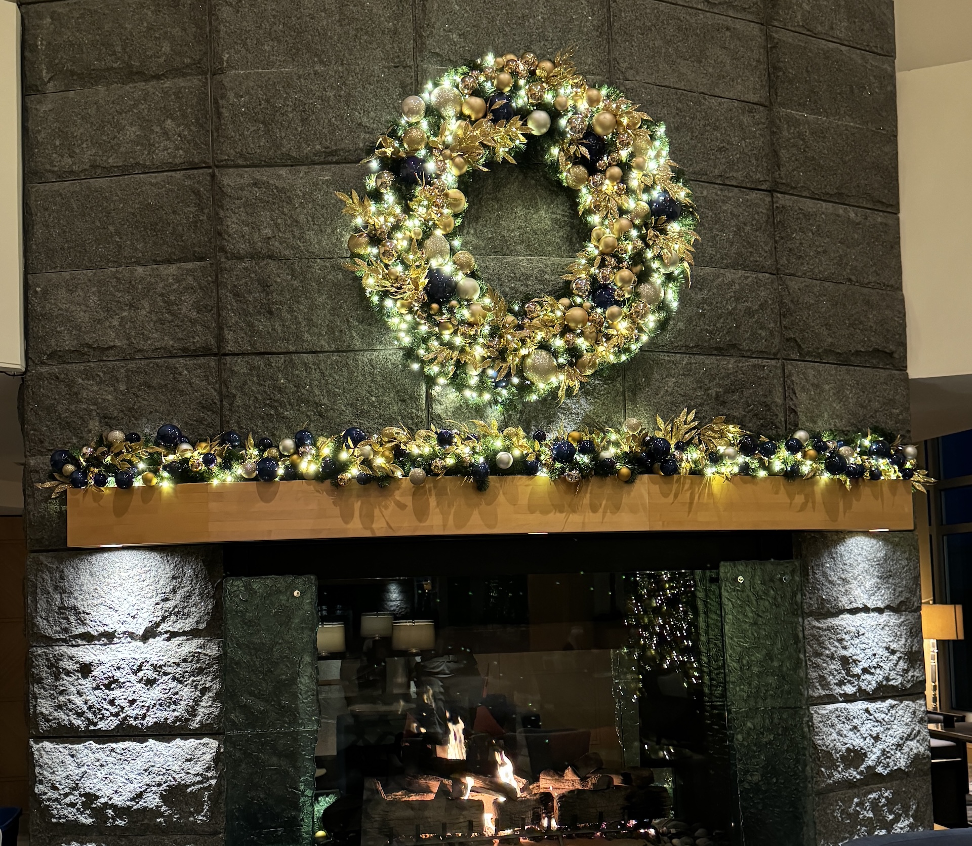 Luxurious Christmas wreath and garland with gold and navy ornaments, warm white lights, and greenery, decorating a stone fireplace in a hotel.