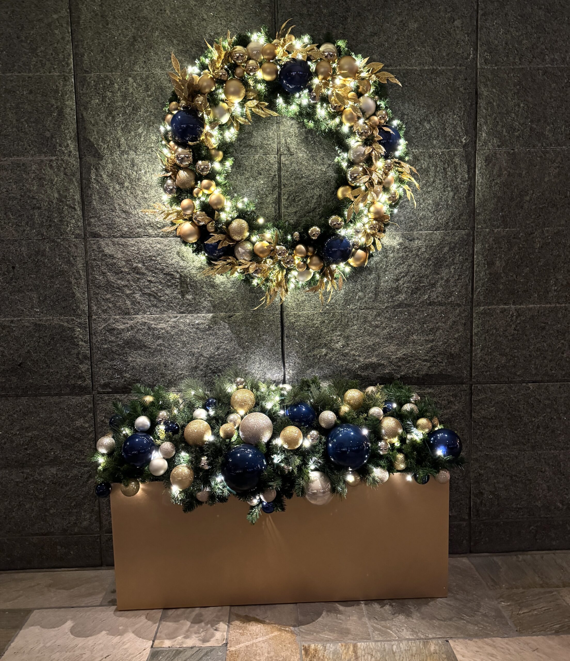 Luxurious Christmas wreath and garland decorated with gold and navy ornaments, warm white lights, and greenery, displayed against a stone wall in a hotel lobby.