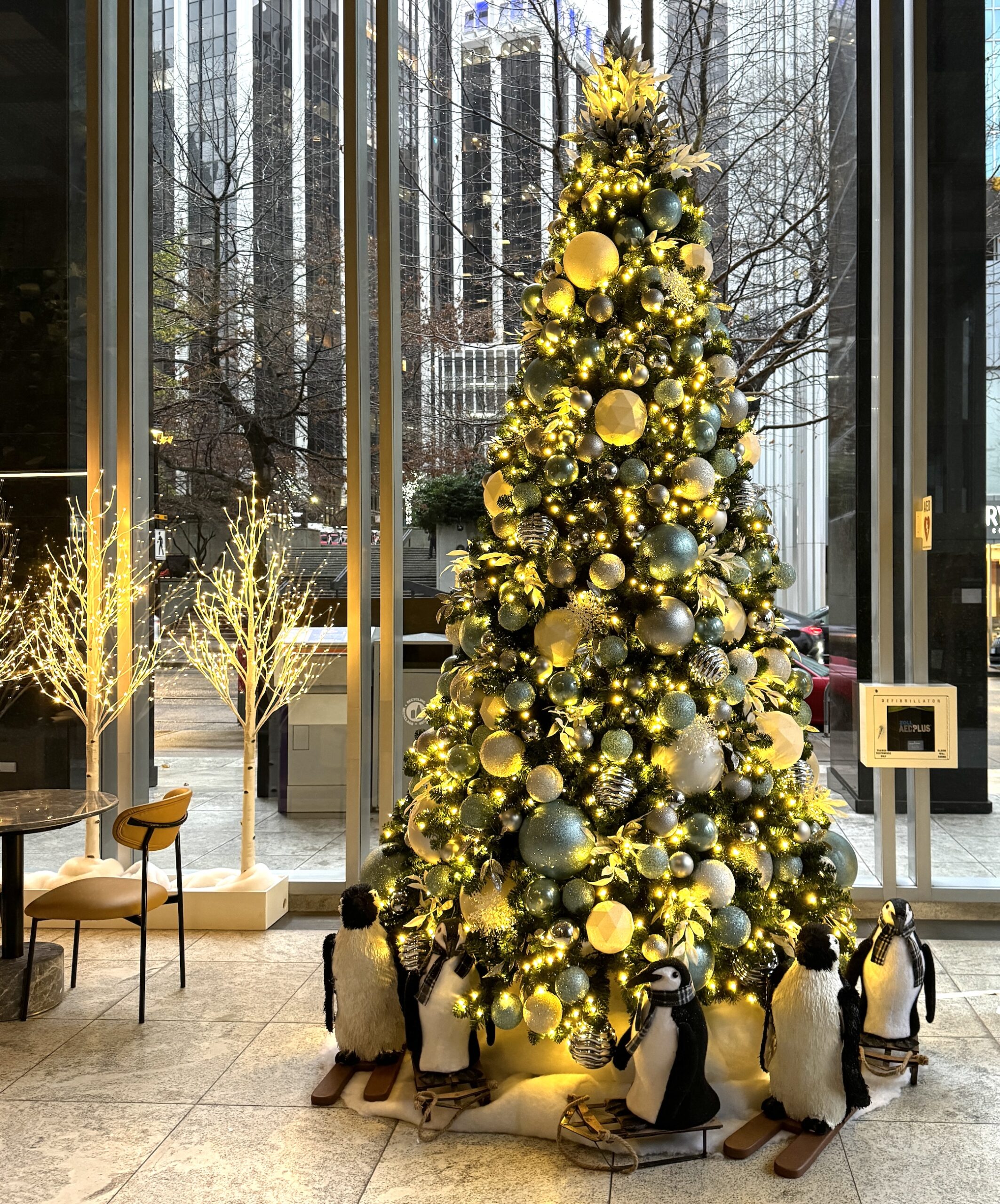 Tall artificial Christmas tree decorated with icy blue, silver, and gold ornaments, warm white lights, and penguin figurines on sleds, set in a modern glass building.