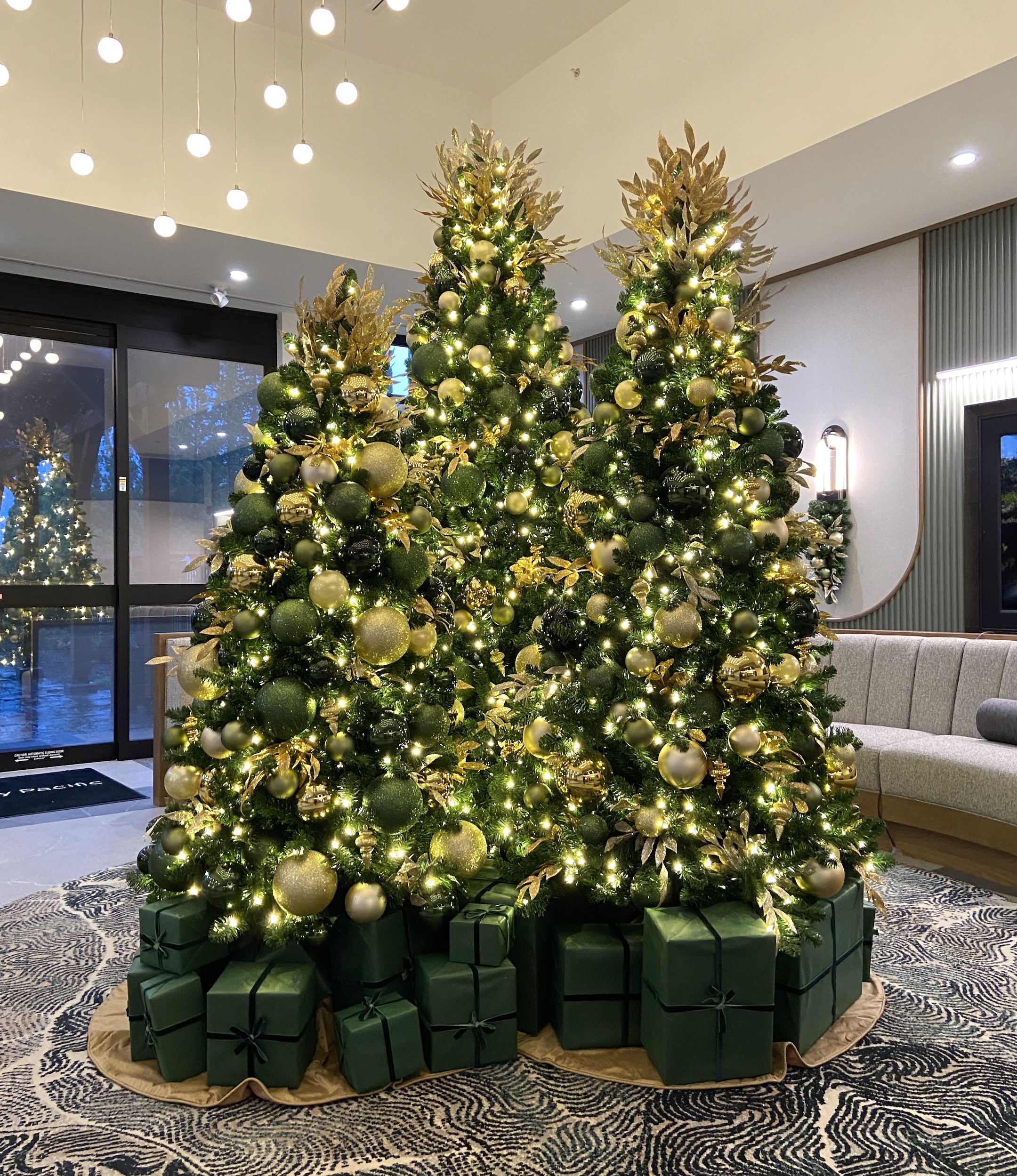 Three stunning Christmas trees decorated in green and gold, surrounded by matching wrapped gift boxes, glowing warmly in a modern resort lobby.