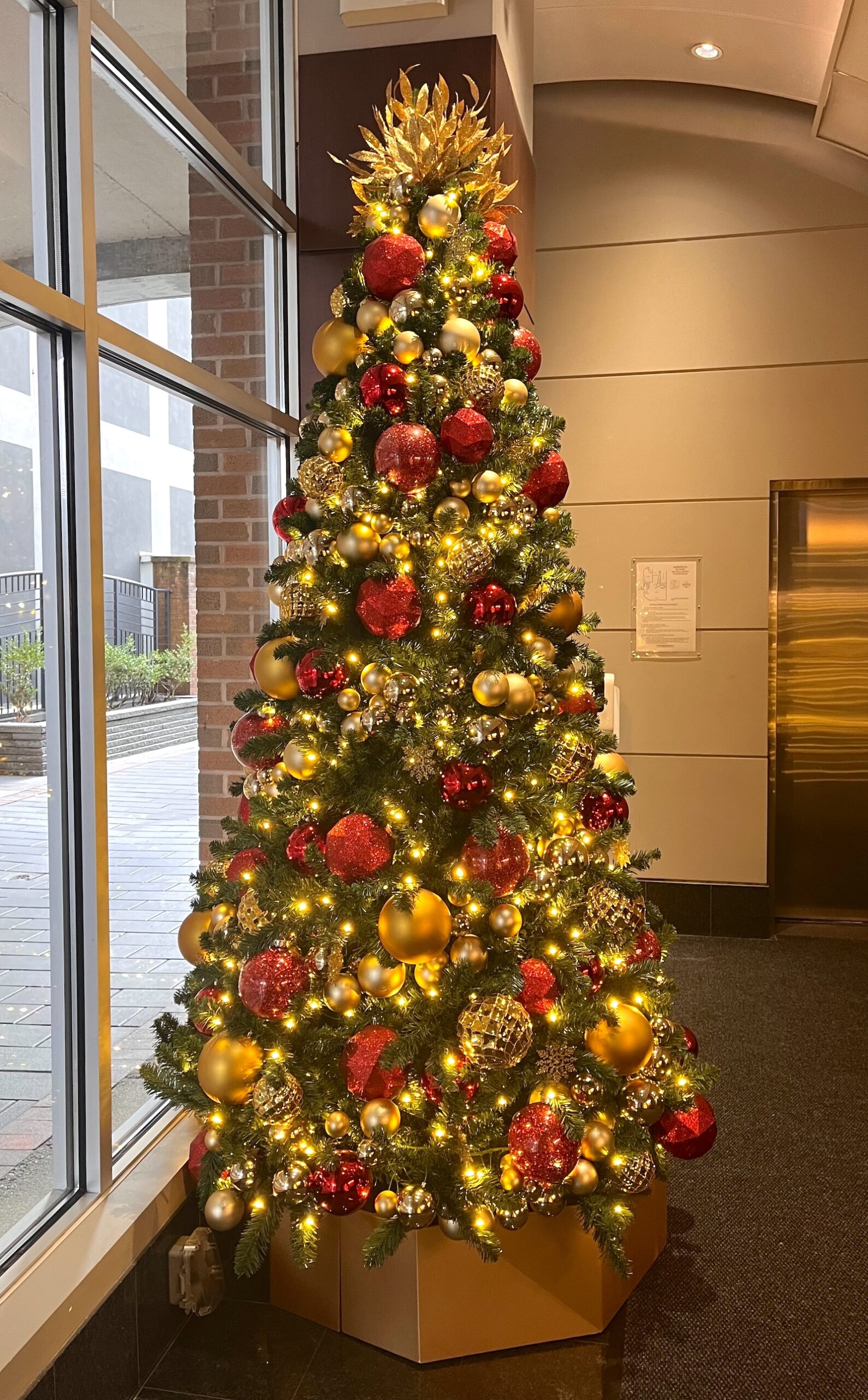Festive Christmas tree decorated with red and gold ornaments, warm white lights, and a golden topper, displayed in a retail office lobby.