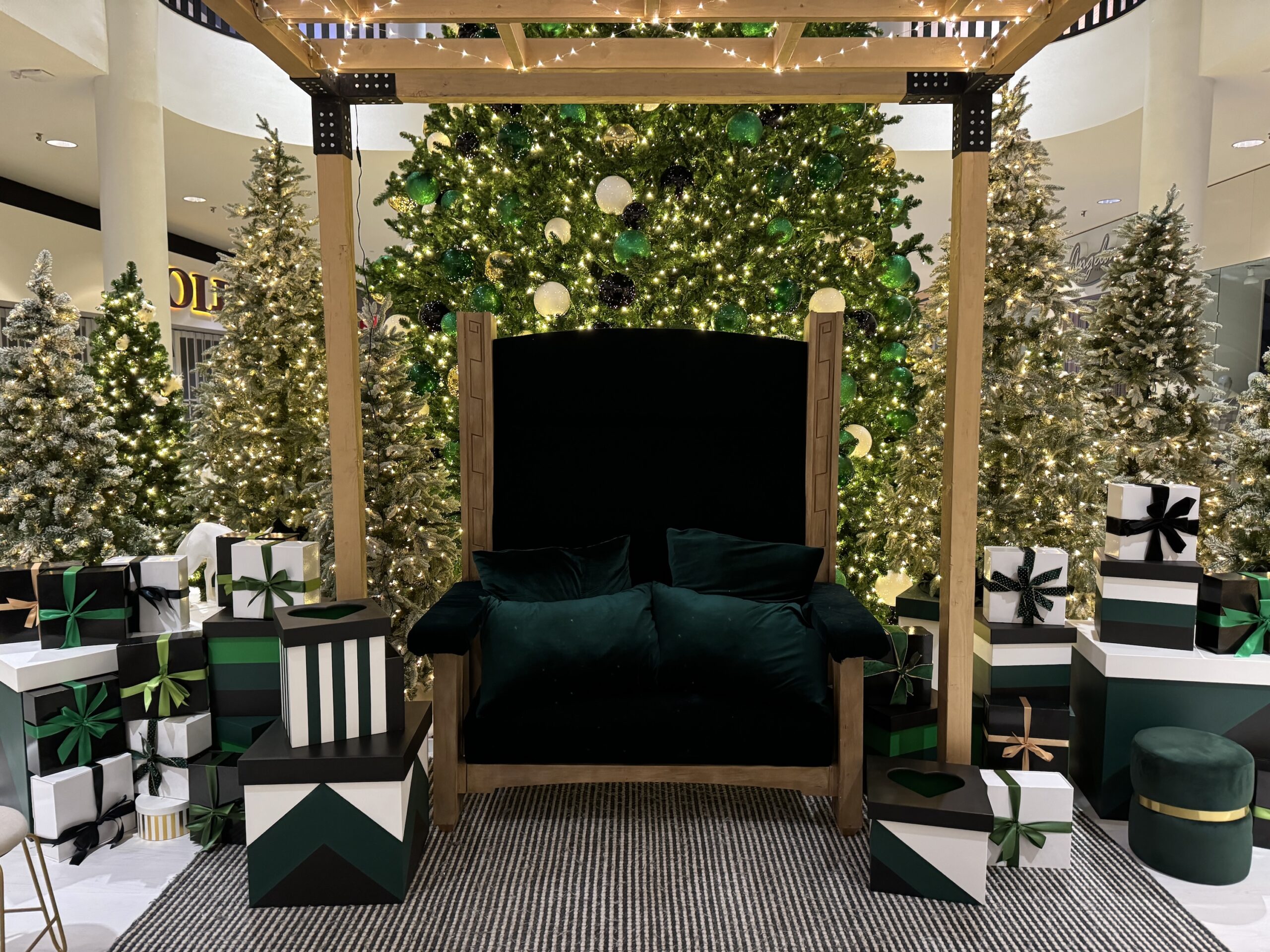 Festive Santa photo op setup featuring green and gold Christmas trees, custom-wrapped gift boxes, and a plush green chair under a lit wooden frame.