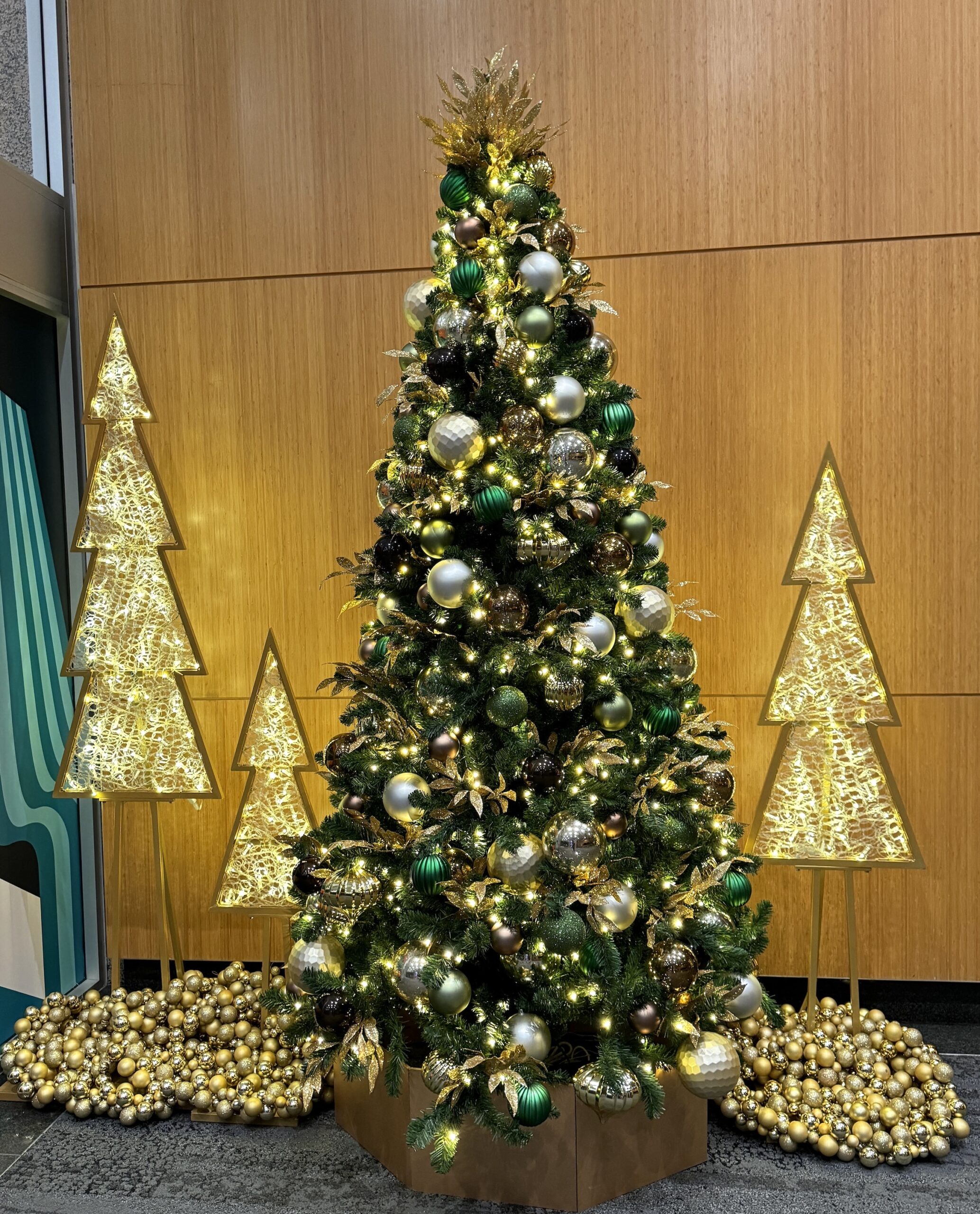 Christmas tree adorned with green, gold, and bronze ornaments, surrounded by illuminated tree sculptures and gold bauble arrangements, displayed in a office lobby in Vancouver.