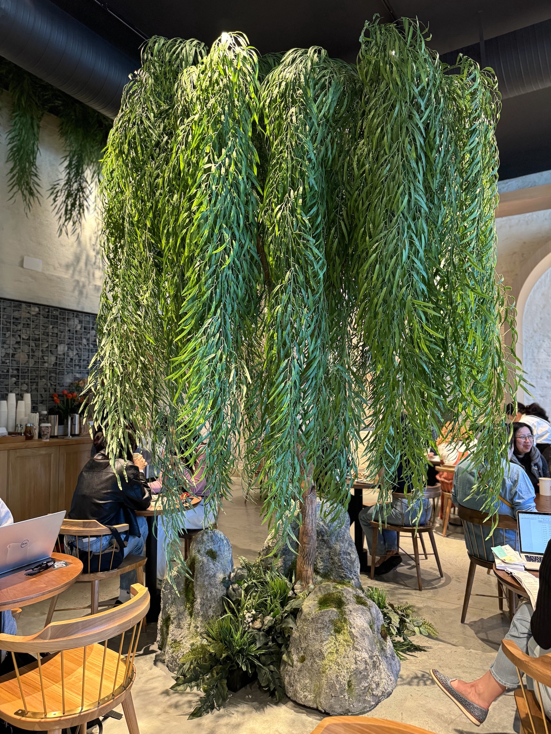 Artificial canopy willow tree with cascading foliage and a rock base in a café. A striking biophilic design feature adding natural ambiance to the modern space.