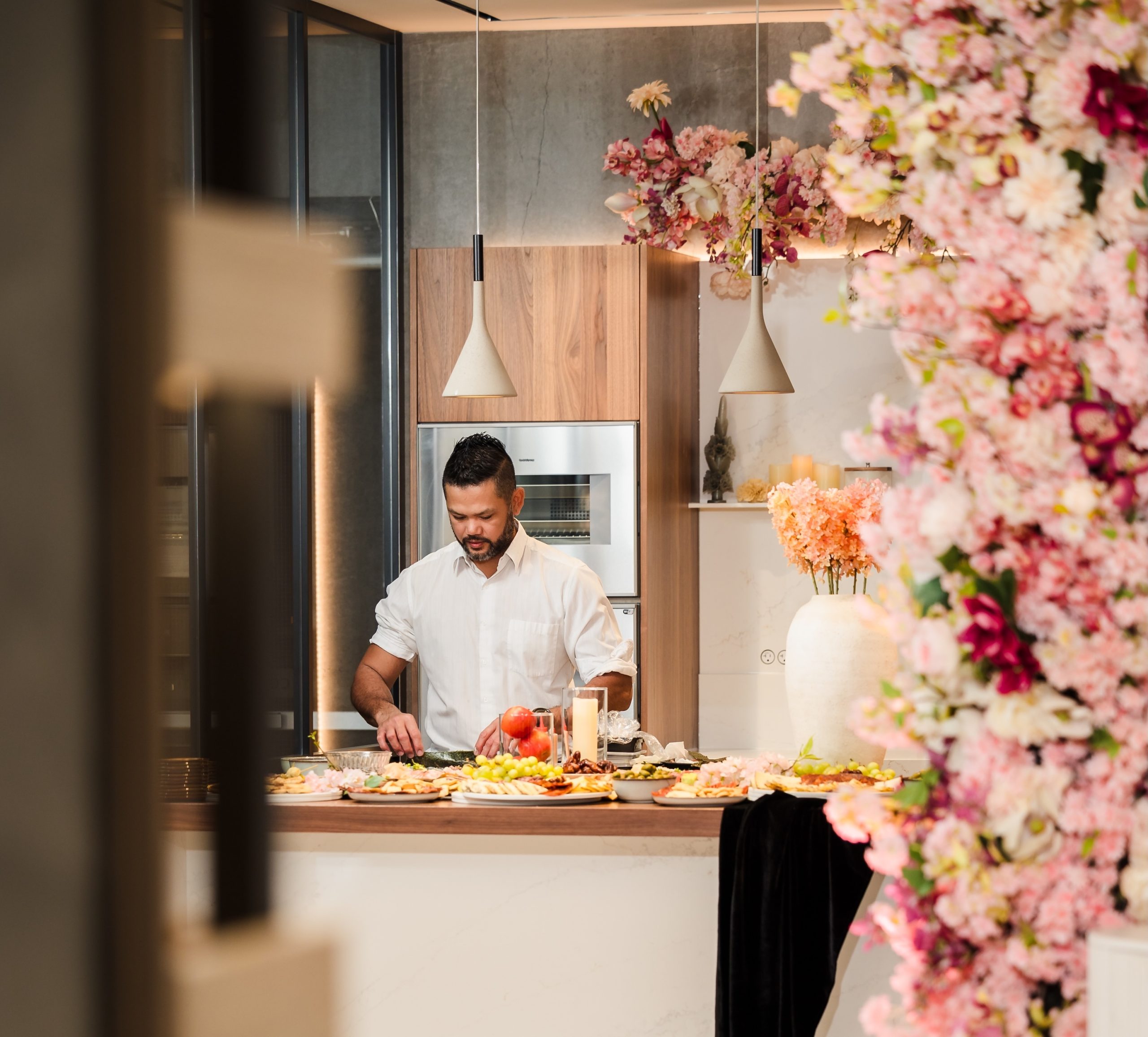 A chef preparing gourmet dishes in a luxury event space, framed by an artificial pink and red floral tower. Elegant event decor.