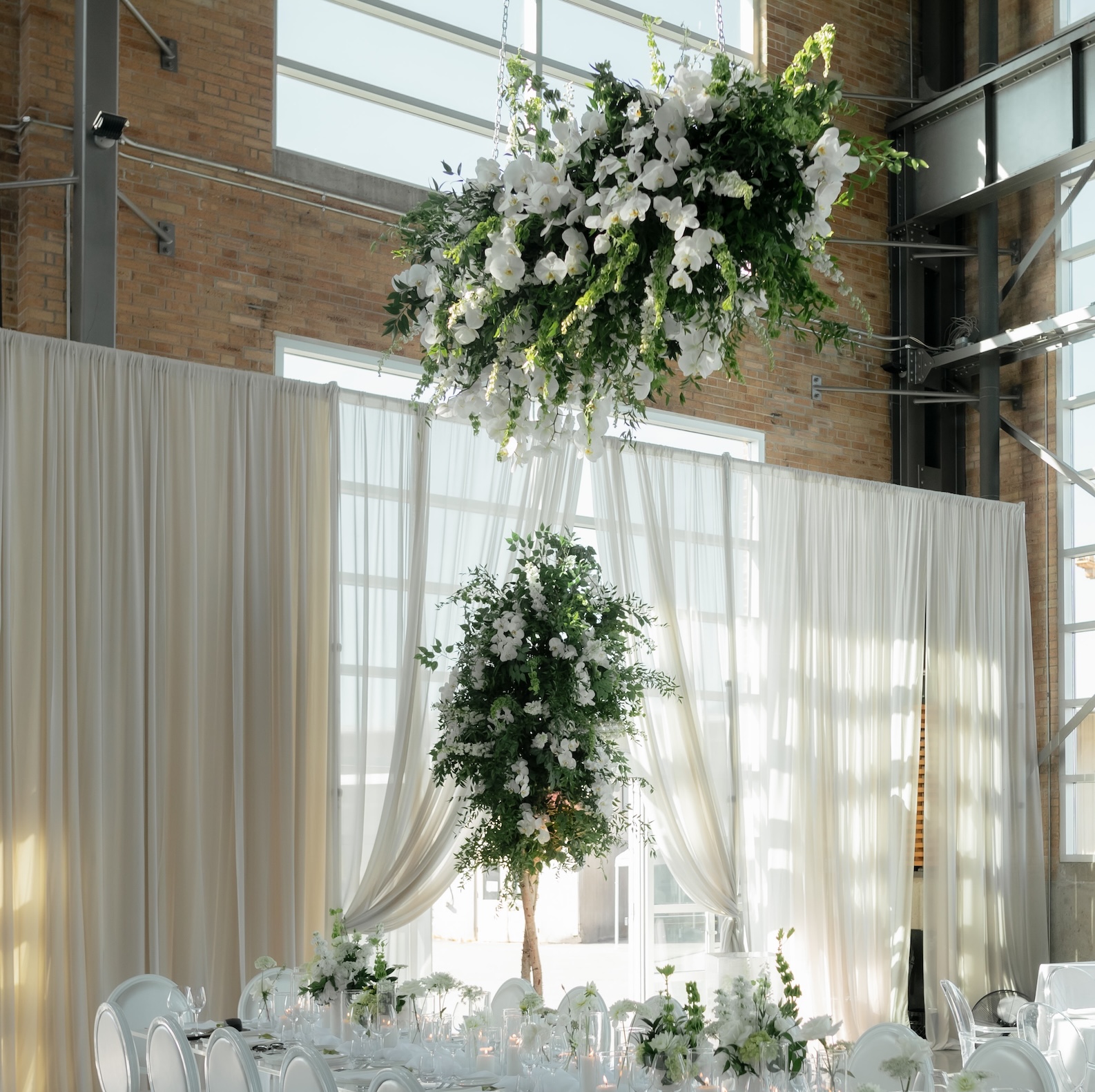 A suspended floral chandelier with cascading white orchids and greenery, paired with a lush artificial tree centerpiece, creating a sophisticated wedding setting.