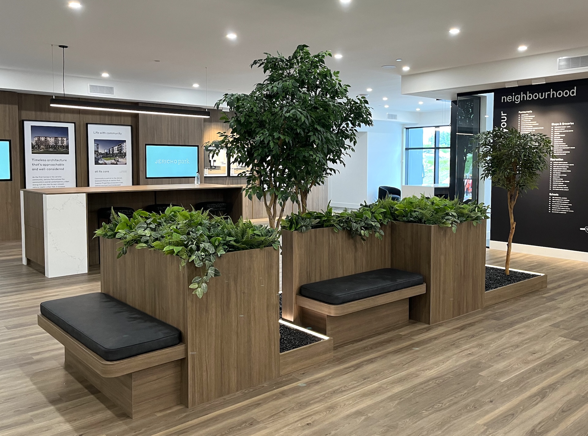 Artificial trees and greenery integrated into wood planters with built-in seating in a modern commercial space. A biophilic design element enhancing the environment.