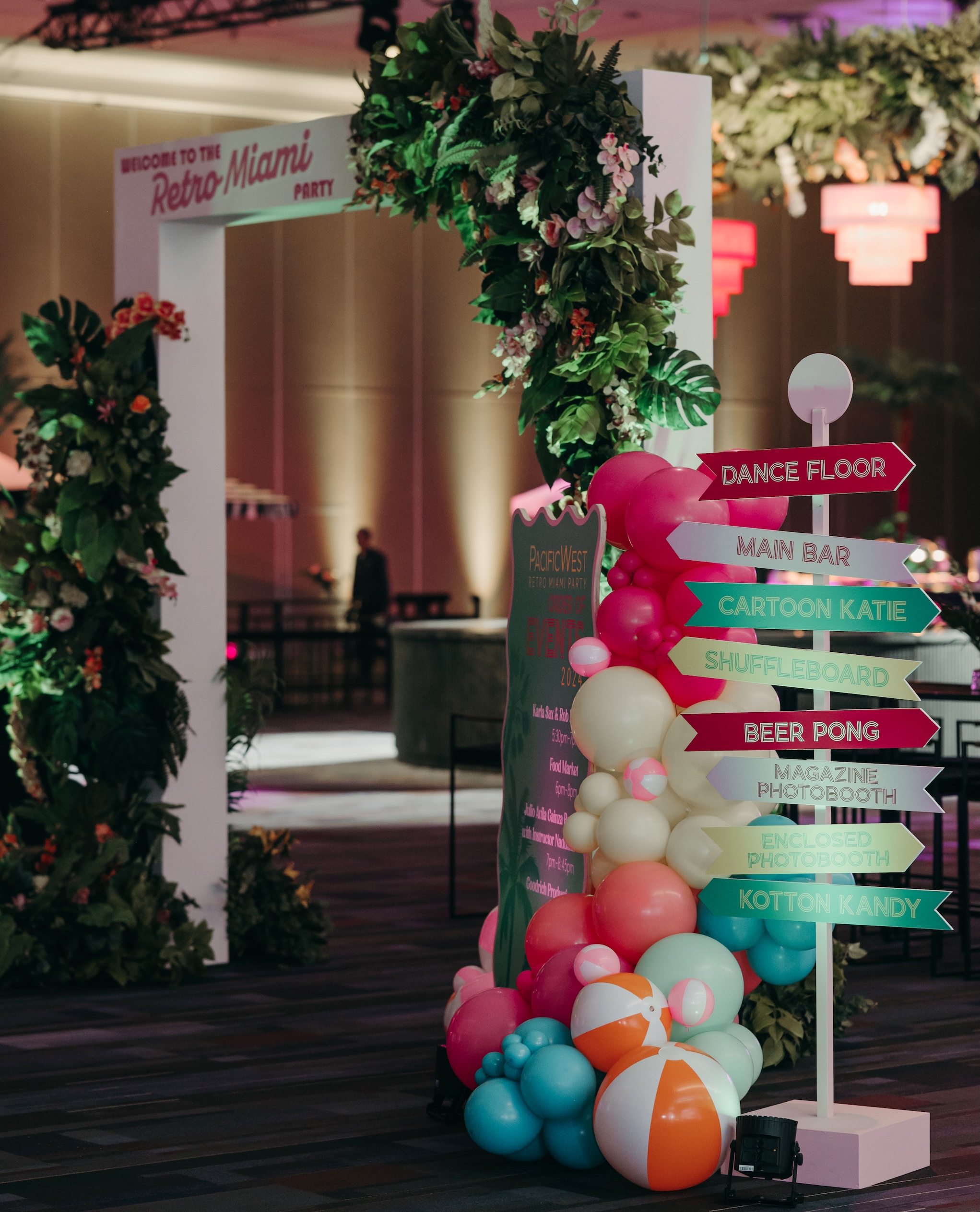 A lively event entrance featuring an artificial tropical floral and greenery arch, colorful balloons, and retro-themed signage.