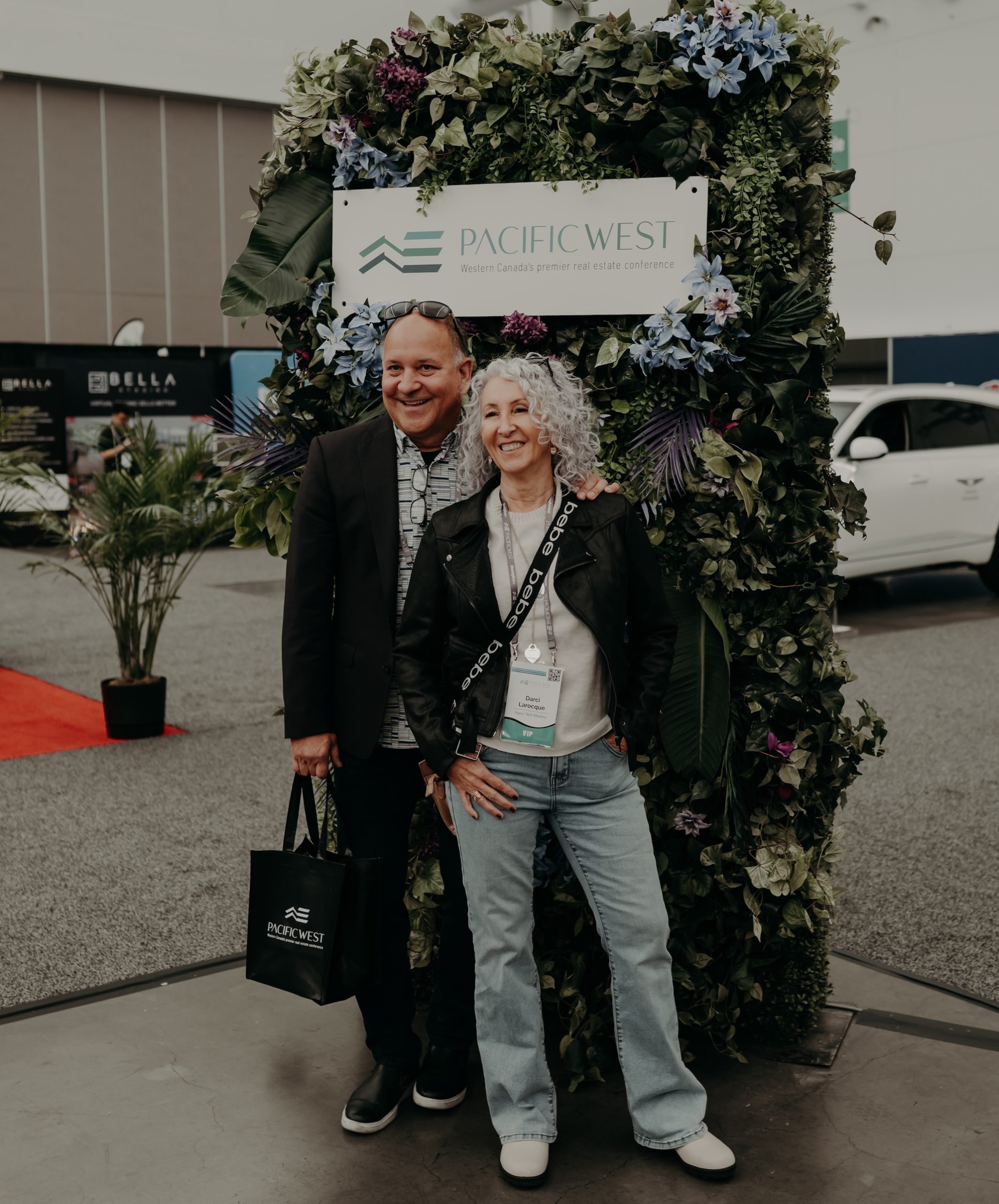 A branded event backdrop featuring artificial tropical flowers, lush greenery, and custom vinyl signage at a corporate conference.