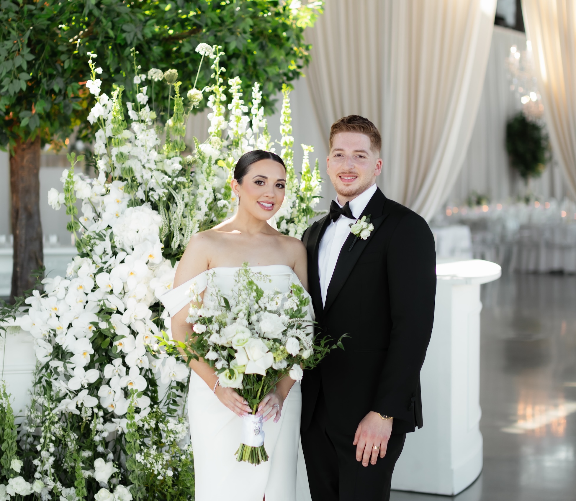 A bride and groom pose beside lush white floral arrangements and an artificial tree in a luxury wedding setting.