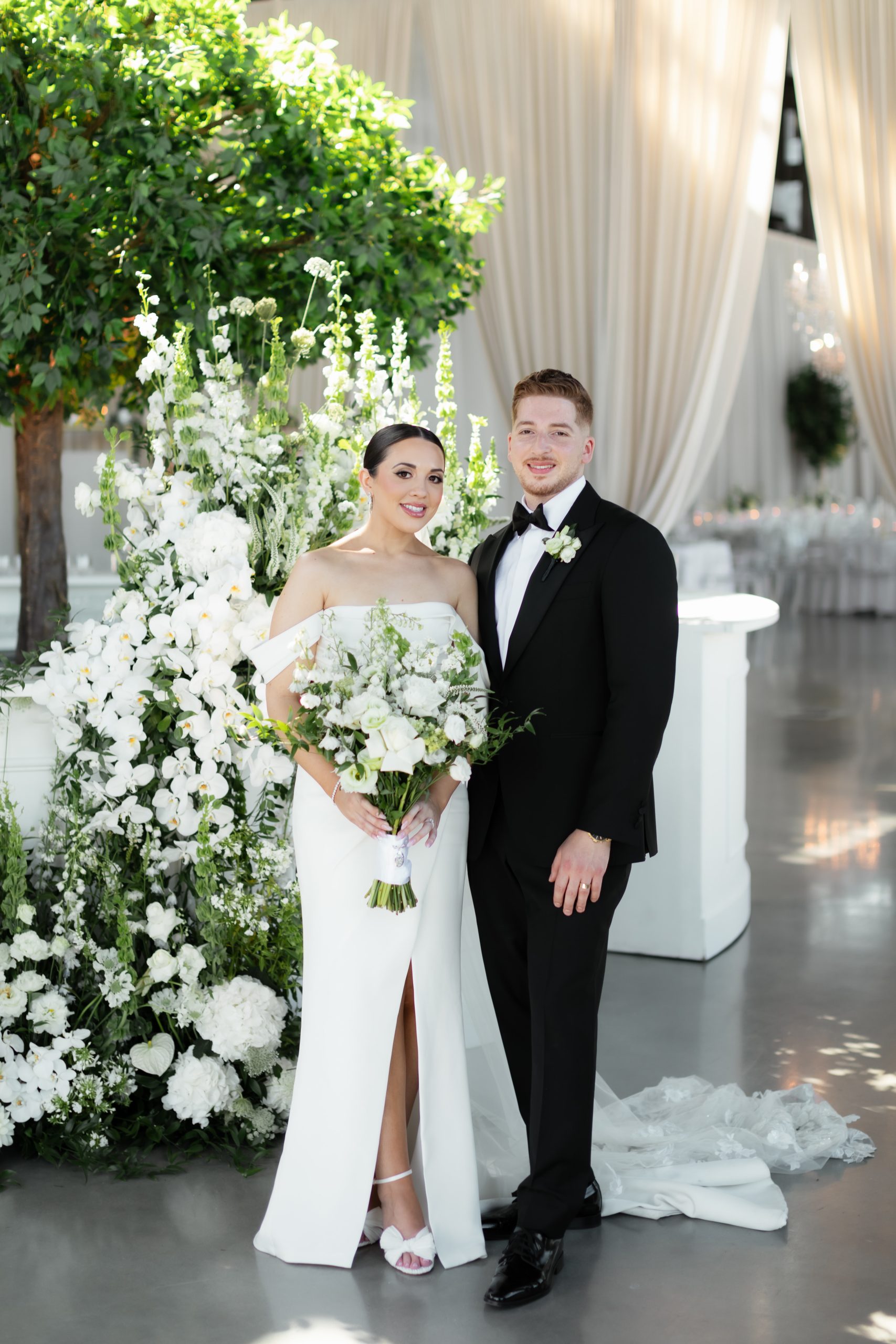 A bride and groom pose beside lush white floral arrangements and an artificial tree in an elegant wedding setting.