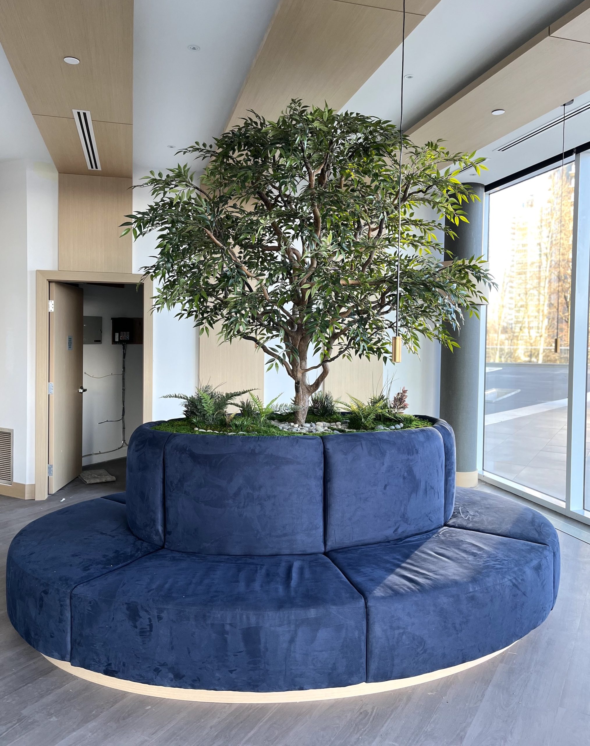 Custom artificial smilax tree in a circular planter with built-in seating. A biophilic design element adding greenery and warmth to a modern interior space.