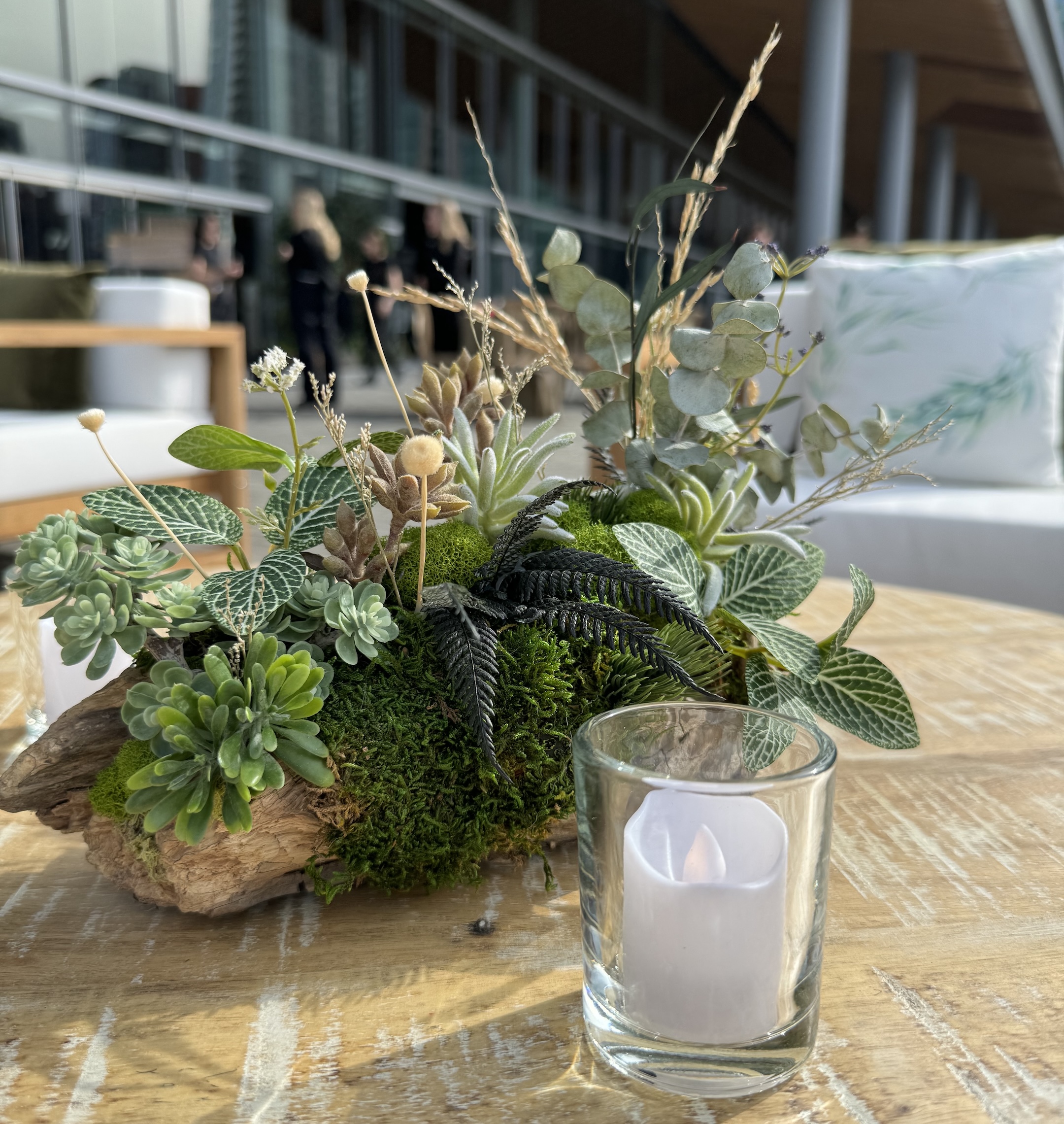 Driftwood centrepiece with faux succulents, moss, and greenery on a wooden table at an outdoor corporate event, with a glass votive candle nearby.