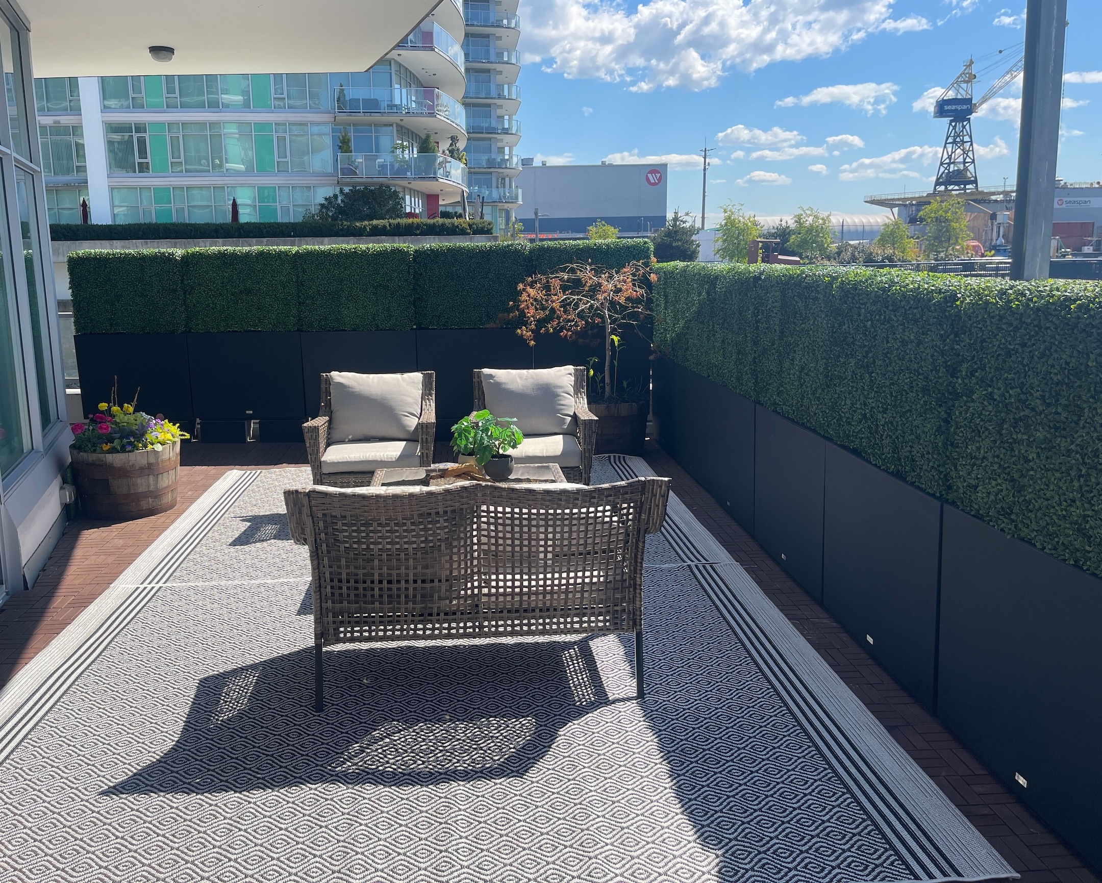 Outdoor patio featuring a fake boxwood hedge privacy wall, woven rattan seating, and a modern black planter setup with city views in the background.