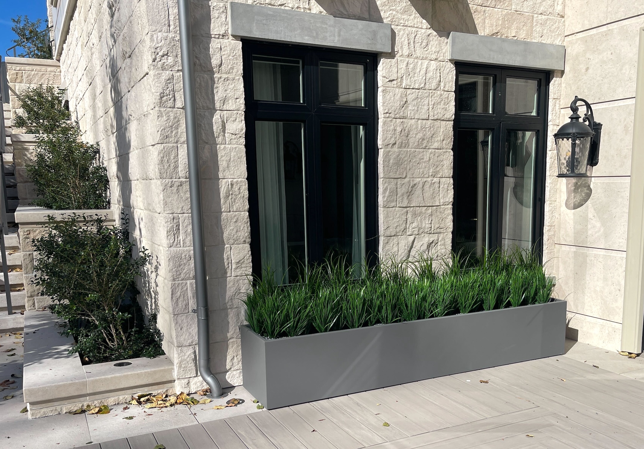 Contemporary outdoor setting featuring a long, grey fake grass planter against a stone-clad building with black window frames.