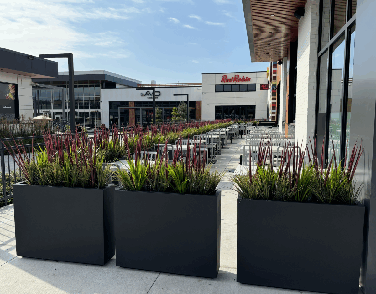 Restaurant patio featuring sleek black planters filled with fake grass and burgundy-tipped greenery, creating a stylish and inviting outdoor dining space.