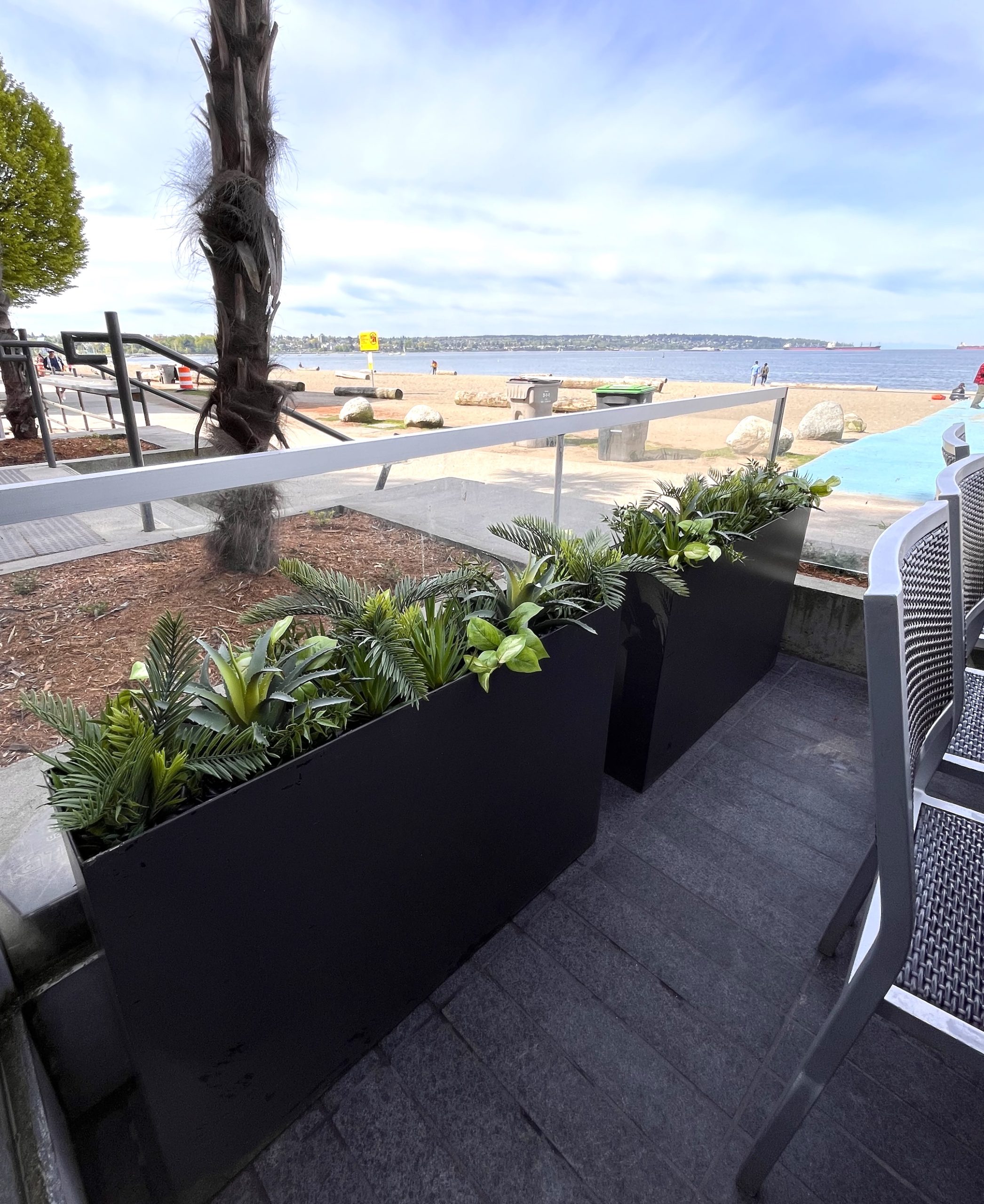 Beachfront patio with sleek black planters filled with faux greenery, offering a natural privacy screen with vibrant foliage.