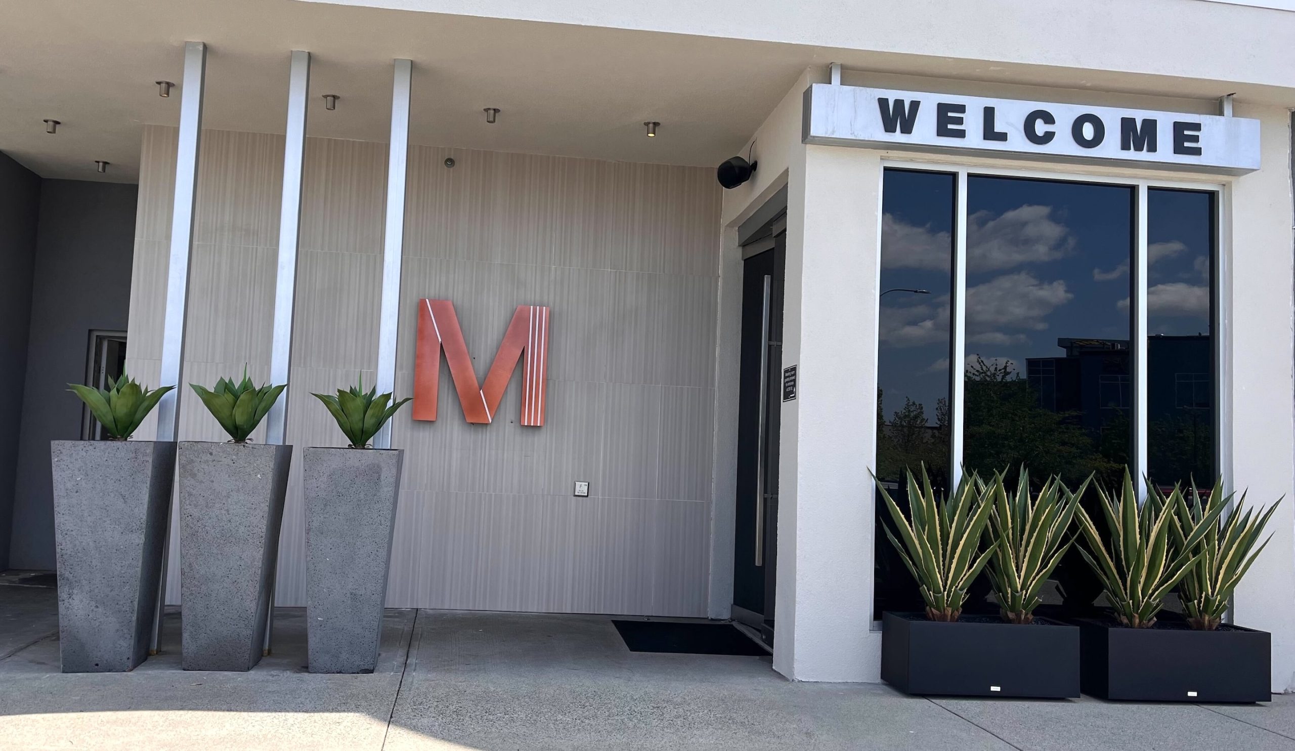 Artificial greenery in modern planters at a restaurant entrance. Features tall concrete and black planters with faux agave plants for a sleek, contemporary look.