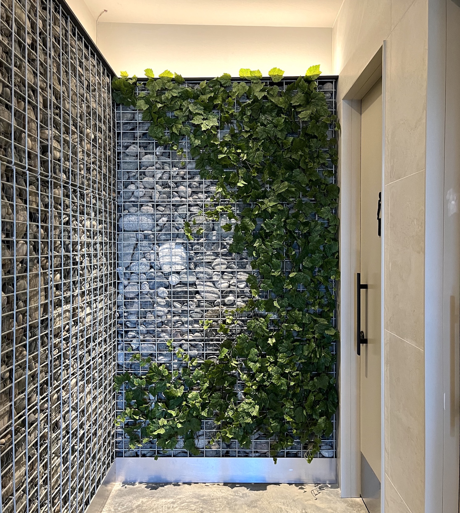 Artificial ivy climbing a gabion stone wall in a modern interior space. A biophilic design feature blending industrial and natural elements for a unique aesthetic.