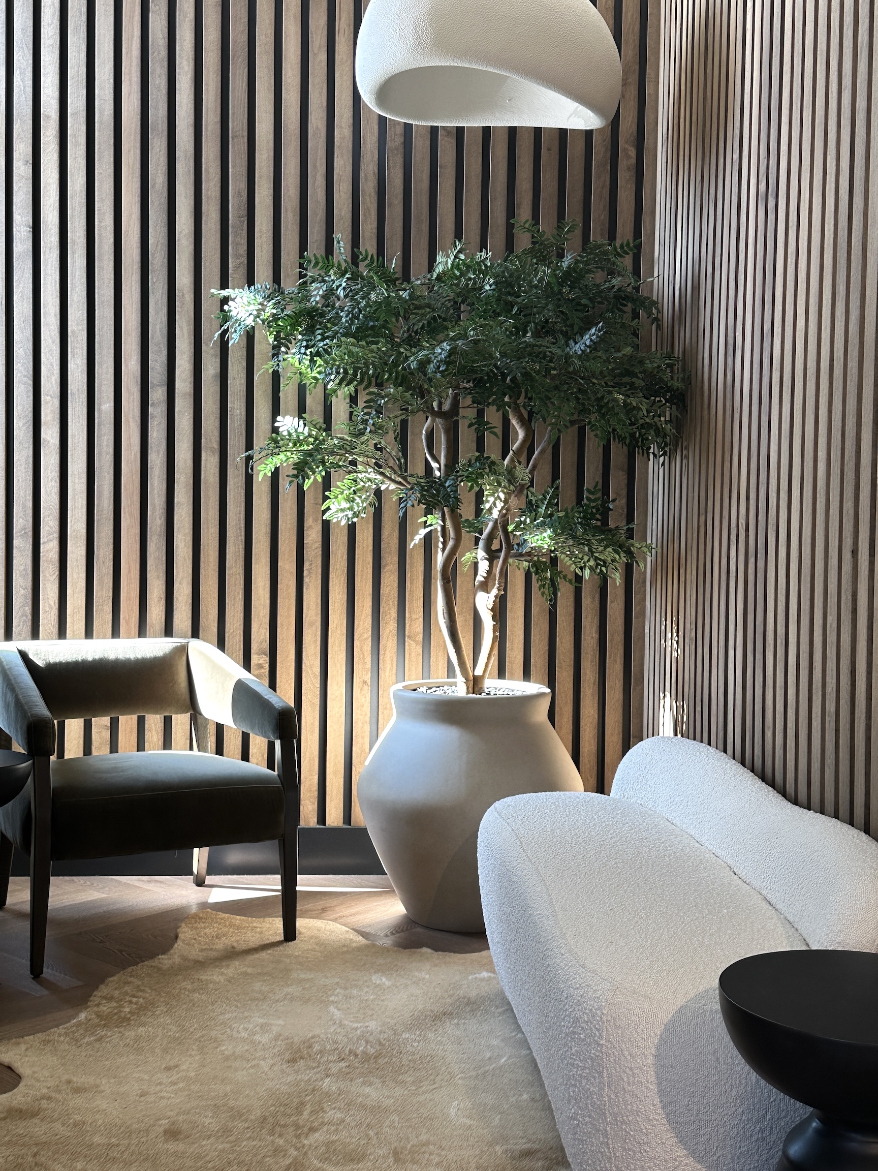 Faux acacia tree in a modern lounge setting. Features a sculptural trunk and lush greenery in a statement planter, complementing wood paneling and soft furnishings.