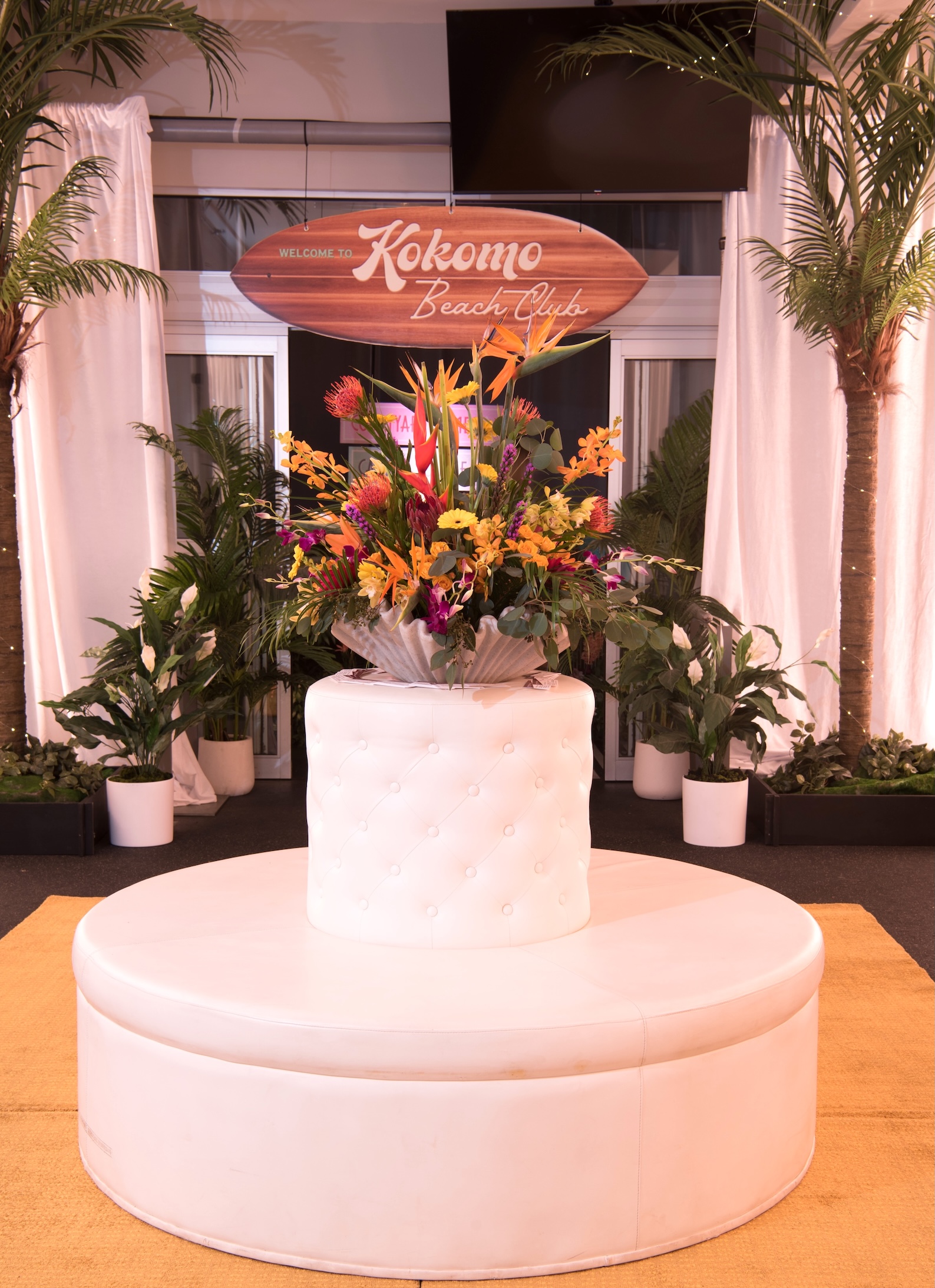 A tropical event setup featuring faux palm trees, lush greenery, and a vibrant floral arrangement in a tufted white lounge display.