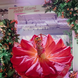 A Miami-themed event entrance with artificial tropical flowers, lush greenery, and a performer in a dramatic floral-inspired costume.