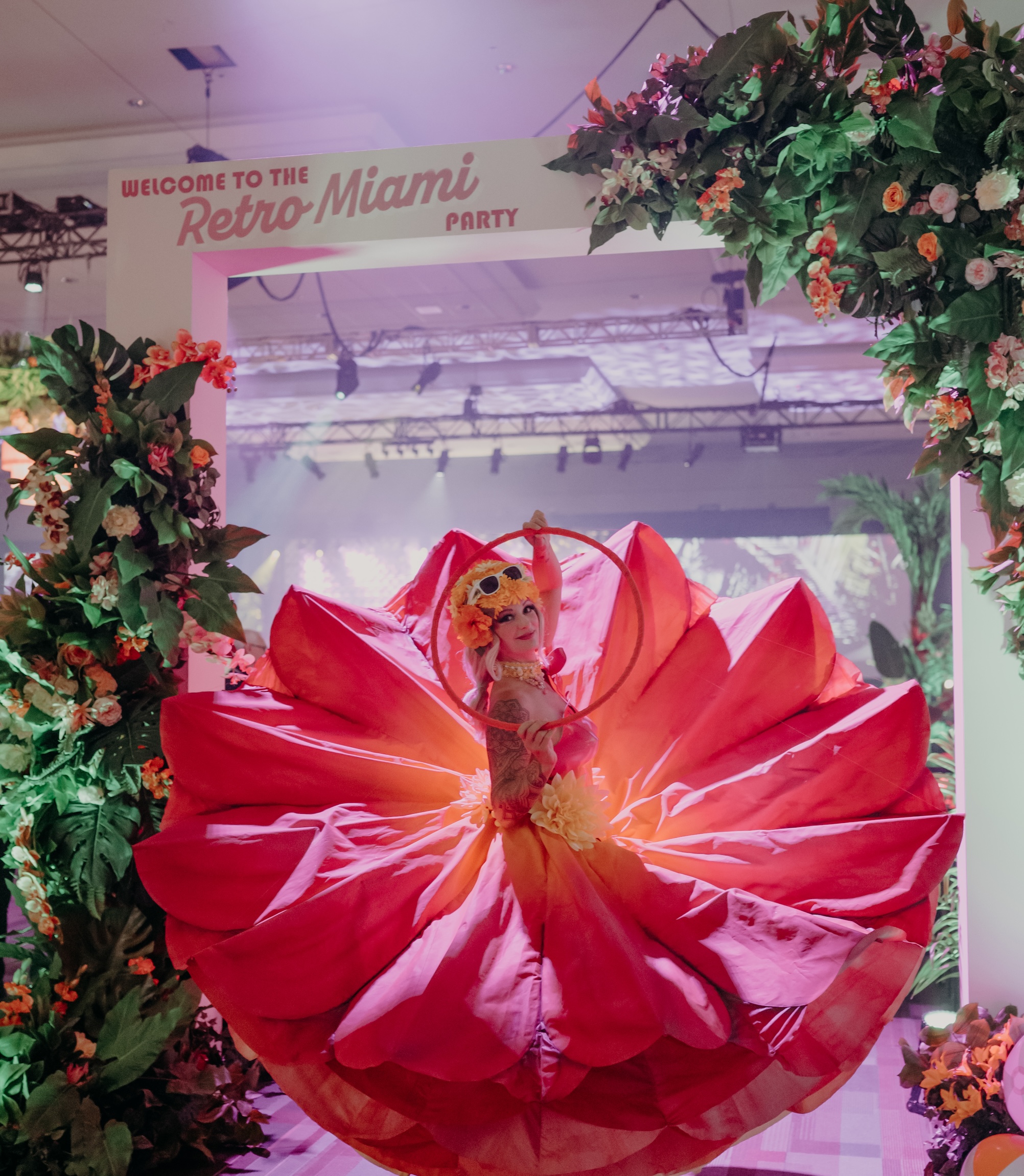 A Miami-themed event entrance with artificial tropical flowers, lush greenery, and a performer in a dramatic floral-inspired costume.