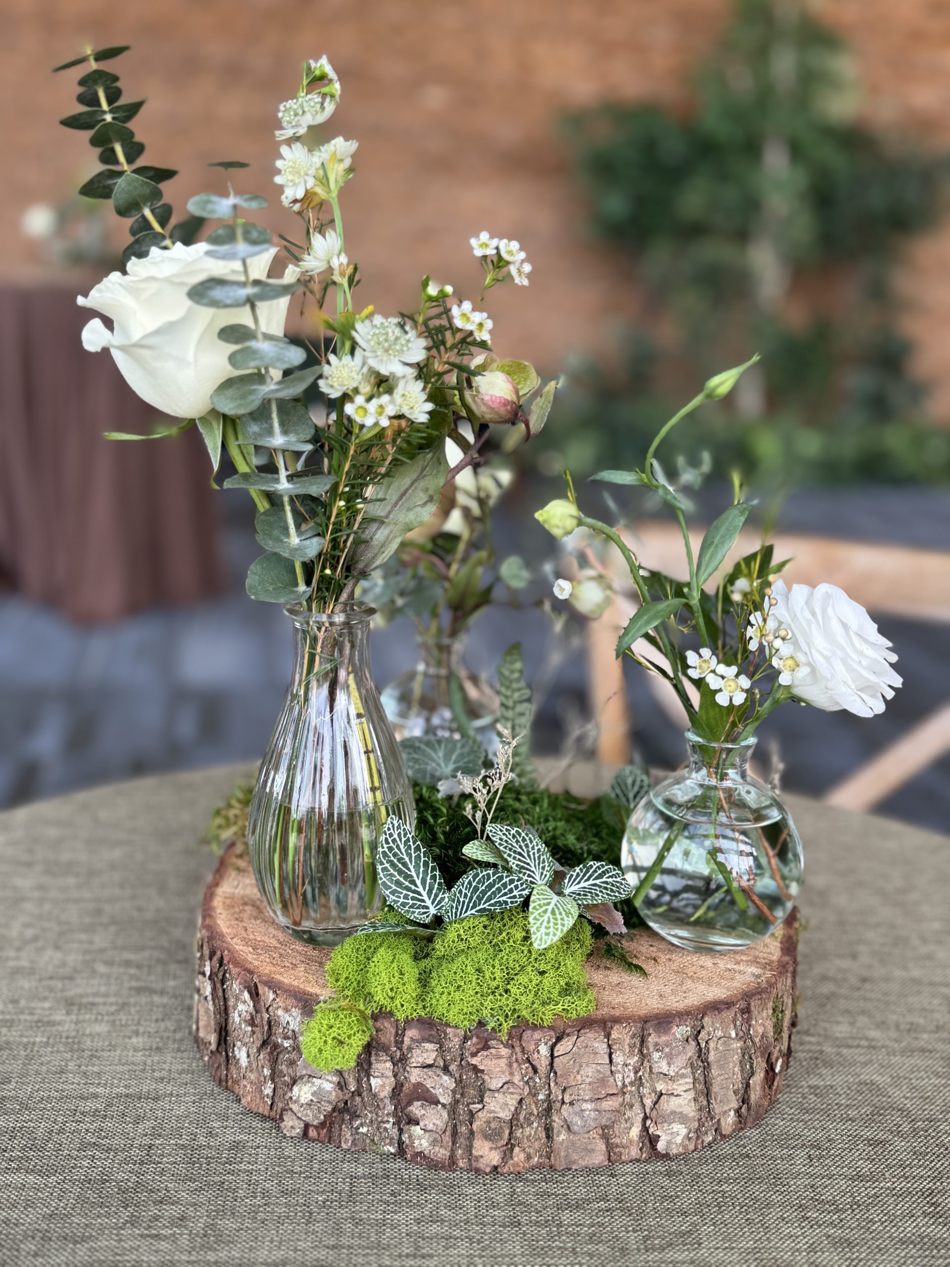 Rustic wood slice centrepiece with moss, greenery, and glass bud vases holding fresh white flowers and eucalyptus at an event.