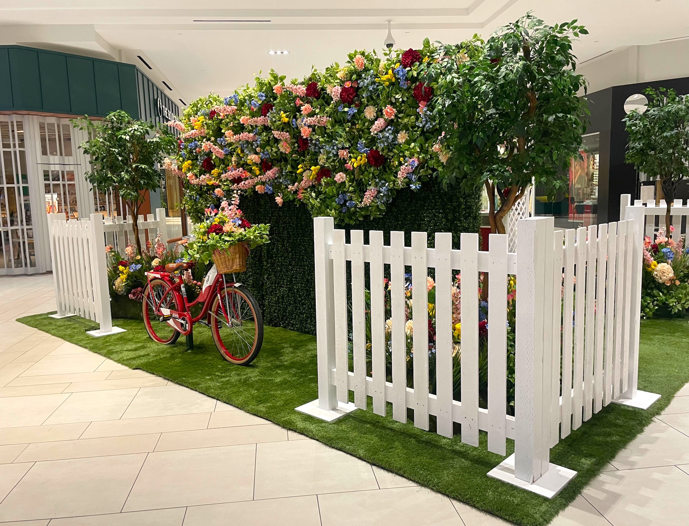 Mother's Day floral photo op display with a red bicycle, vibrant flowers, a greenery wall, white picket fences, and lush trees in a Langley shopping center.