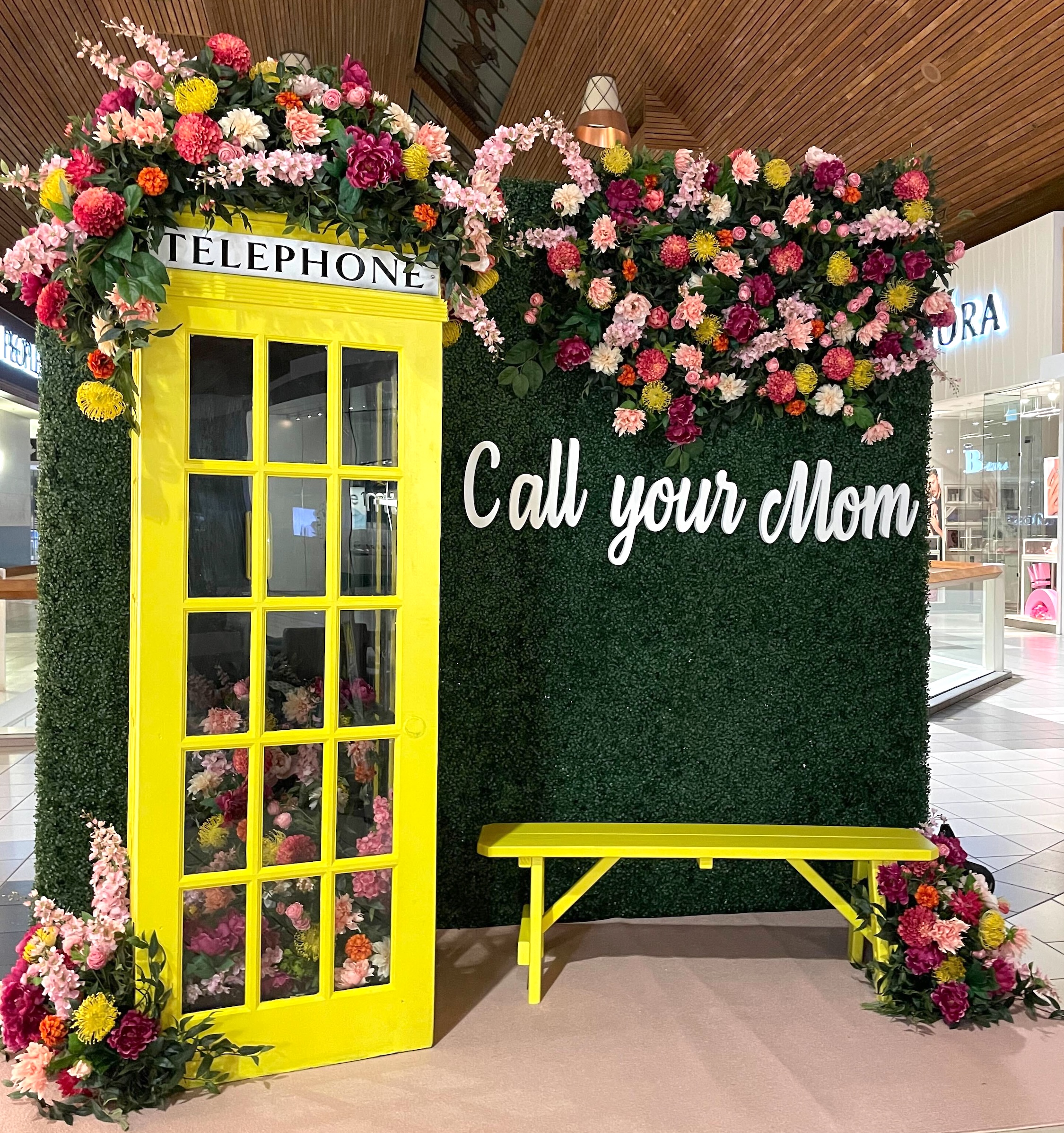 Mother's Day photo op display with a yellow telephone booth adorned with colorful artificial flowers, an artificial green hedge wall, and a "Call your Mom" sign in a shopping centre.