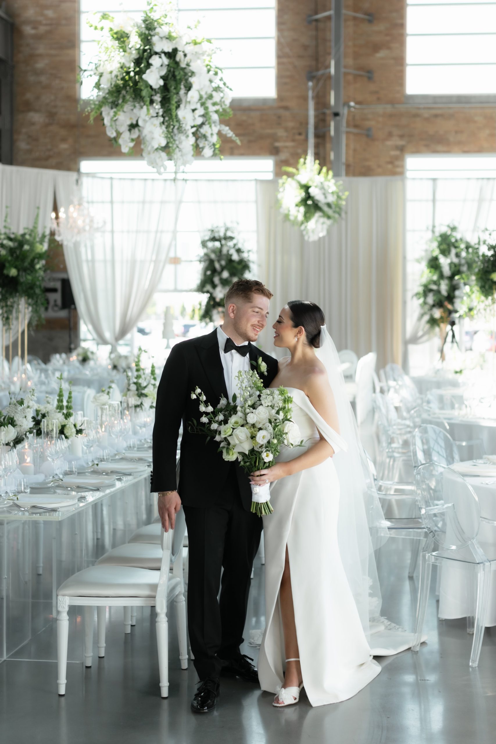 A bride and groom share a moment in a wedding reception space with floral chandeliers, elegant greenery, and modern white décor.